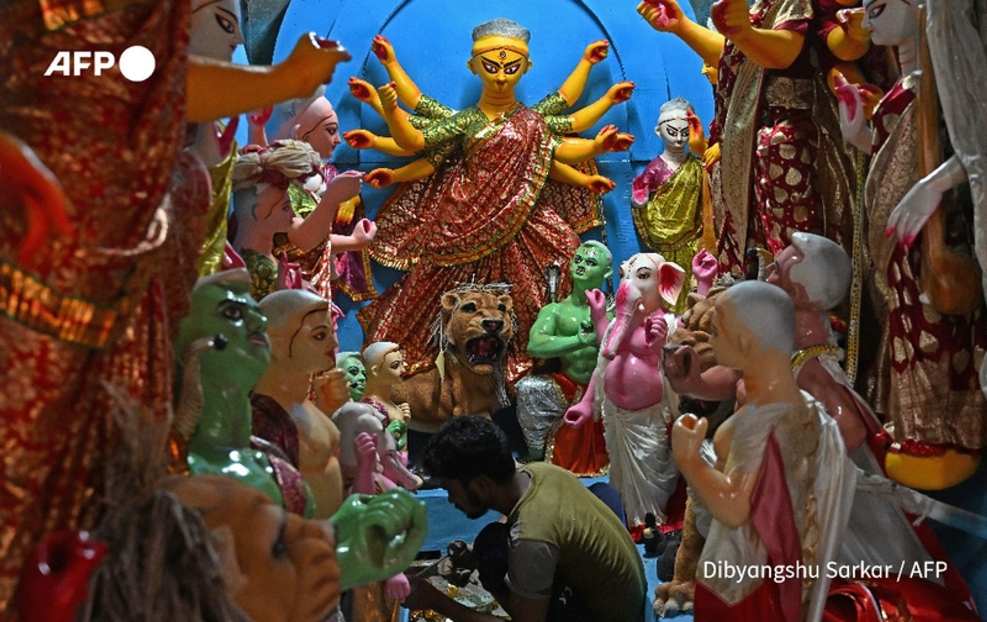 A vibrant scene depicting a Hindu ceremonial setting, centered around a large idol of the goddess Durga. She is adorned in ornate clothing, with multiple arms outstretched, each holding symbolic objects. The goddess has a striking expression with bright, piercing eyes and is seated on a lion, representing strength. Surrounding her are smaller idols of various deities and figures, all intricately designed with vivid colors, displaying expressions of devotion. In the foreground, a man is kneeling and working meticulously, possibly preparing the idols or the area for a ritual. The backdrop is painted in bright blue, enhancing the festive atmosphere. The overall scene exudes a sense of spirituality and reverence, reflecting the cultural significance of this ritual in Kolkata.