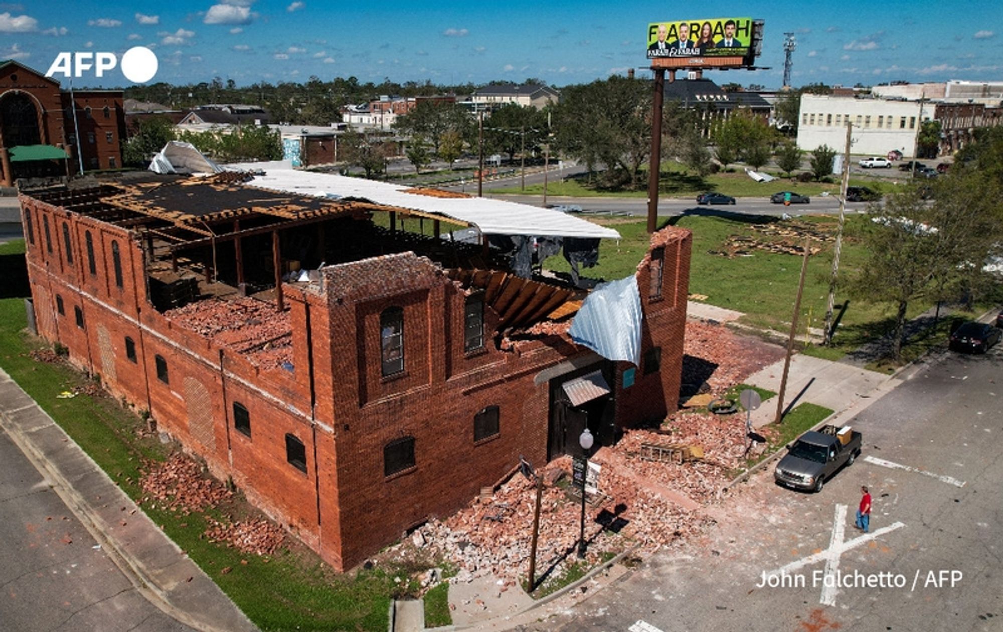 A scene showing significant damage to a red brick building, which appears to have lost its roof and suffered extensive structural damage. Debris is scattered around the site, including fallen bricks and pieces of the roof on the ground. A visibly distressed man is seen walking near the building, indicating the impact of the storm. In the background, there are additional buildings and trees under a clear blue sky, suggesting a calmer atmosphere following the chaotic weather. A large billboard can be seen in the upper corner, displaying the word "FAIRBRANCH" with additional advertising details below. The image captures the aftermath of storm Helene, reflecting the devastation in the area, where the death toll has risen, and many households remain without power.