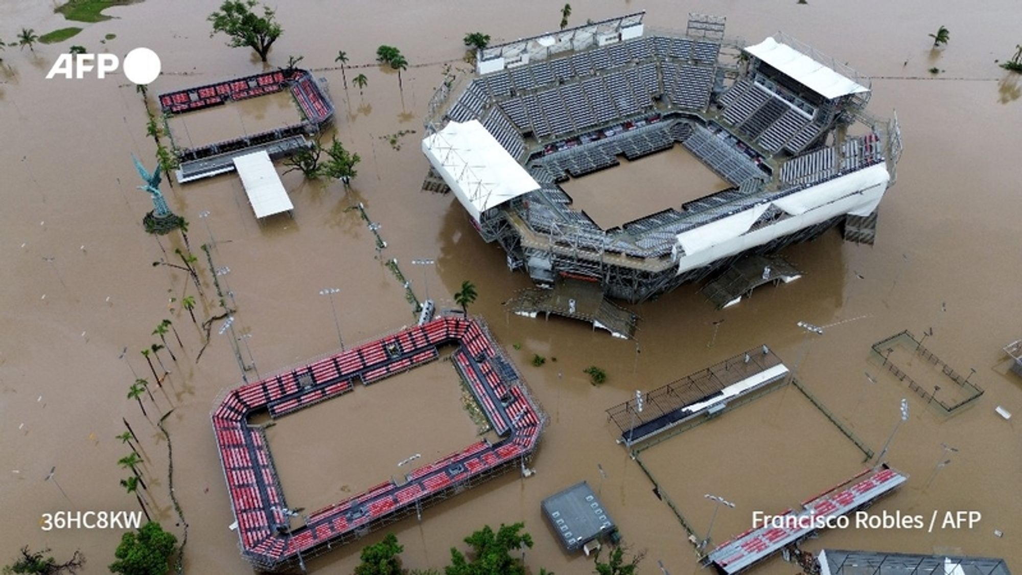 Aerial view of a large stadium and surrounding sports fields in a flooded area, reflecting the impact of Hurricane John in Acapulco, Guerrero, Mexico. The stadium features a partially collapsed grandstand, with empty rows of seating partially submerged under brown water. Around the stadium, two rectangular sports fields, also flooded, can be seen, with the outlines of the playing surfaces visible. Several tall palm trees rise above the water, indicating the flood level. The sky is overcast, suggesting a somber atmosphere, and debris is scattered throughout the flooded area, underscoring the destructive effects of the hurricane. This scene highlights the severe damage and ongoing recovery challenges facing the locality after the storm.