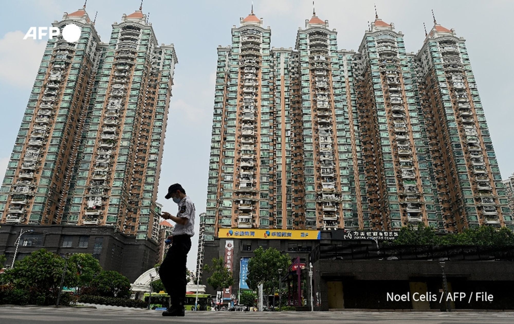 A security guard stands on a walkway in the foreground, holding a smartphone and looking at the screen. He is dressed in a white shirt with black trousers and a black cap, and is positioned in front of several tall residential buildings. In the background, five high-rise apartment complexes tower, showcasing a modern architectural style with a mix of glass and concrete. The buildings feature a green and beige color palette, with balconies and window designs that indicate urban living. Below the buildings, a commercial area is visible, with signs partially obscured but suggesting retail and services. The sky appears partly cloudy, revealing a muted blue backdrop. The scene conveys a sense of urban life amid China's ongoing housing market developments.