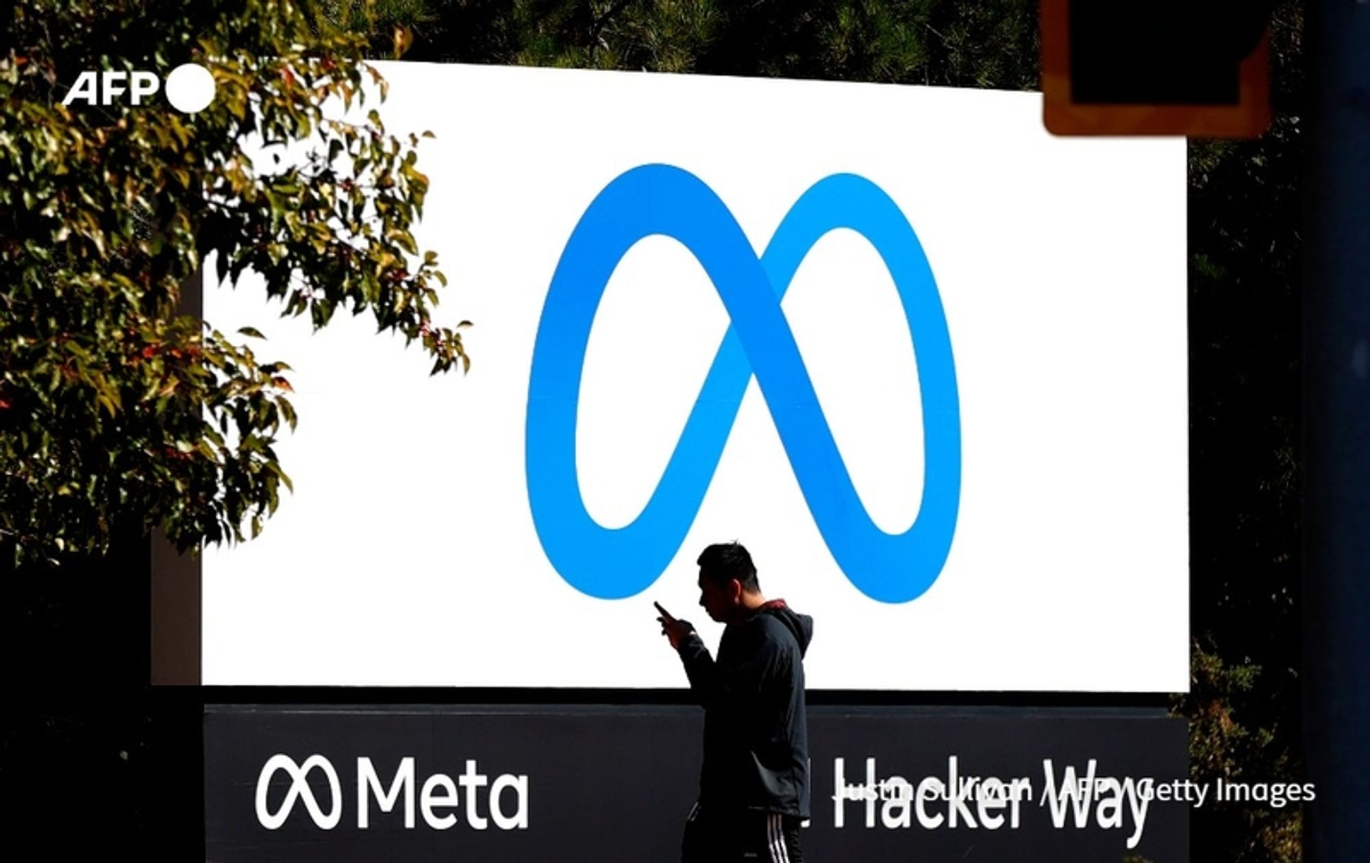 A person stands on a sidewalk in front of a large advertisement for Meta, the parent company of Facebook. The individual is looking at their phone, dressed in a dark hoodie and jeans. In the background, there is a prominent white sign displaying the Meta logo, which consists of an abstract infinity symbol in shades of blue. Below the logo, the word "Meta" is clearly visible in a bold, black font, situated above the words "Hacker Way," which also appear in black. The surroundings include some lush green foliage, suggesting a well-maintained environment around the corporate office. The light conditions indicate a bright day. The scene highlights the intersection of technology and daily life.