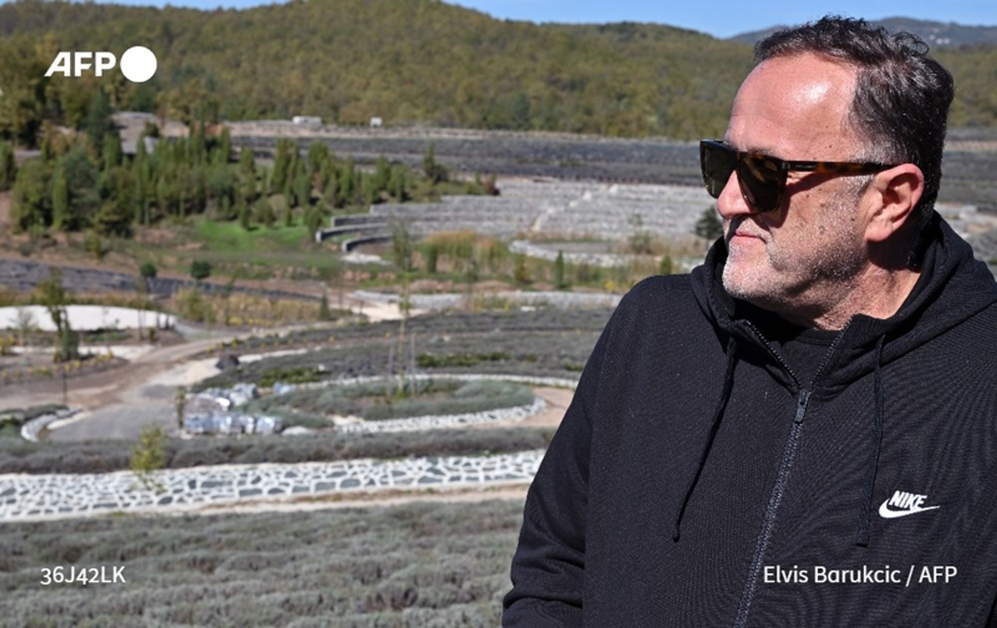 A man stands in the foreground, wearing a black zip-up jacket with a hood and sunglasses. He has short, dark hair and a beard. Behind him, a sprawling landscape of a garden is visible, designed to resemble Vincent Van Gogh's painting "Starry Night." The garden features intricate patterns formed by rows of plants, with a variety of greenery creating flowing curves. The area is terraced, with small trees and shrubs scattered throughout. A hill with dense vegetation rises in the background, while the sky is clear, suggesting a sunny day. The overall scene is a vibrant blend of nature and artistry, reflecting the creativity of the individual who designed this unique tribute to Van Gogh's art.