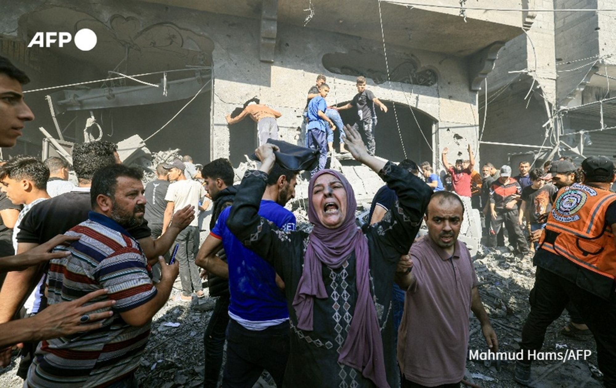 A chaotic scene in an urban area after a recent bombing. The foreground shows a group of people reacting to the devastation, with expressions of shock and distress. A woman wearing a dark hijab, with a patterned shirt underneath, stands out as she raises her hands in despair, her mouth open as if screaming. To her left, a man in a striped shirt appears to be reaching out towards her. Surrounding them, various individuals are in motion; some are helping others who may be injured while others are climbing over debris. The background features a partially collapsed building with exposed concrete and scattered rubble, indicating the severity of the damage. A rescue worker in a bright orange vest can be seen assisting in the response efforts. The atmosphere conveys a sense of urgency and the human impact of conflict, emphasizing the need for reports on such crises. The image is attributed to Mahmud Hams, a photographer for AFP.