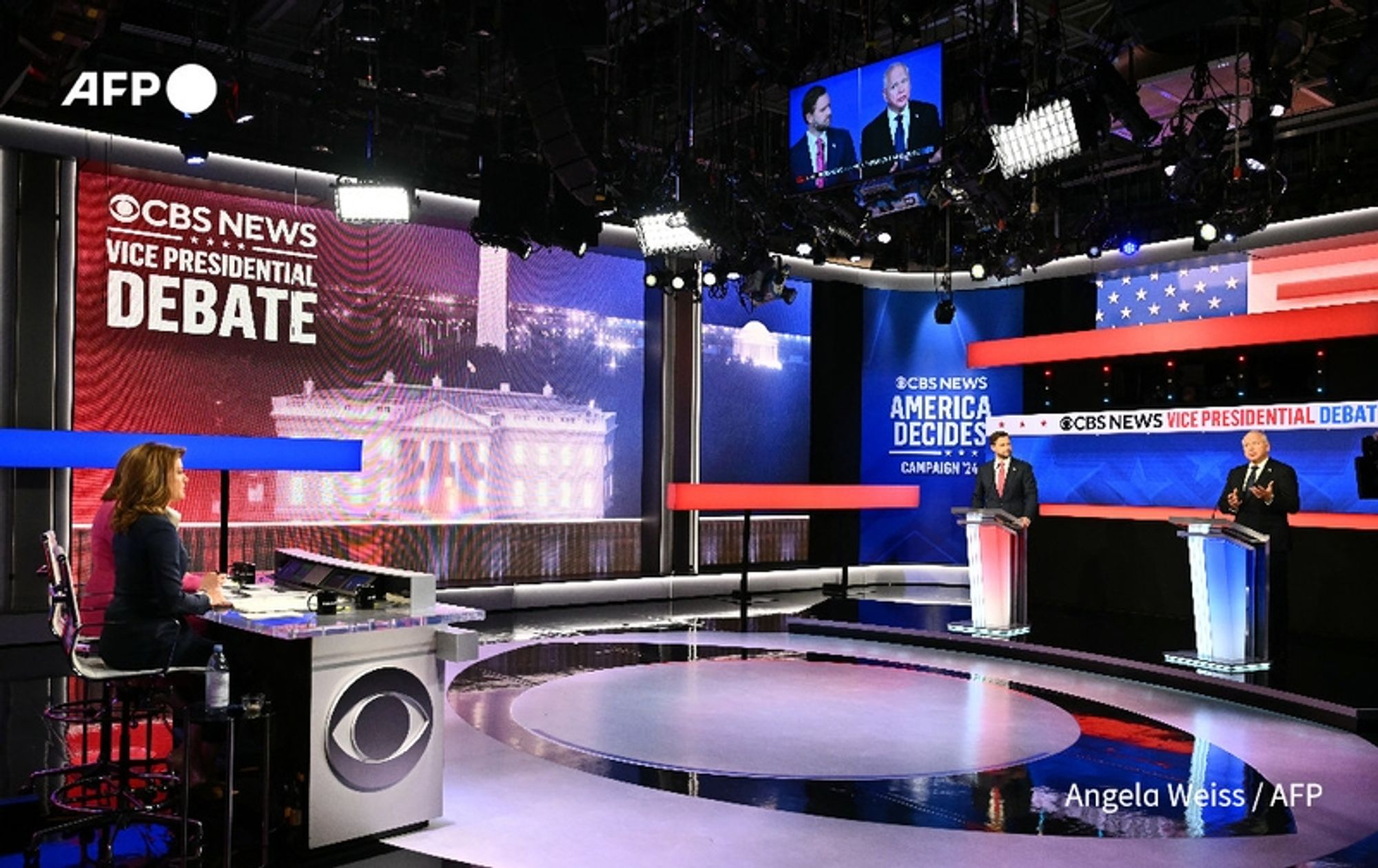 A television studio set for a vice presidential debate, featuring two candidates, J.D. Vance and Tim Walz, standing behind separate podiums. The backdrop includes large screens displaying "CBS NEWS AMERICA DECIDES" and "CBS NEWS VICE PRESIDENTIAL DEBATE." The stage is brightly lit, with red, white, and blue elements, reflecting the theme of an American political event. A female moderator, seated at a sleek desk with a CBS logo, is visible on the left side of the image, attentively observing the debate. Above the candidates, a screen shows images of both participants during the discussion. The floor has a circular design, enhancing the studio's modern look. The overall atmosphere conveys a high-stakes political event, with a focus on swing state dynamics in the upcoming elections.