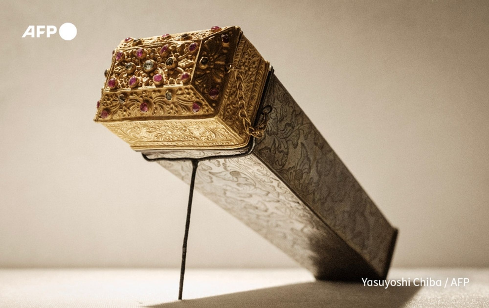 A detailed close-up of a beautifully crafted jewelry artifact from the Lombok collection, displayed prominently at a repatriation ceremony. The object is primarily elongated and box-shaped, featuring an ornate lid covered in intricate gold detailing, with flush, colorful gemstones including rubies and sapphires embedded in a decorative pattern at the top. The lid has an angular, triangular design, emphasizing the careful craftsmanship. The body of the jewelry box has a rich, dark finish, adorned with swirling engravings that add to its artistic value. The artifact is mounted on a slender stand, allowing it to be viewed from multiple angles, against a softly lit background that highlights its luxurious materials and intricate details. This artifact signifies cultural heritage and artistic achievement, displayed in the context of a significant event at the National Museum of Indonesia in Jakarta.