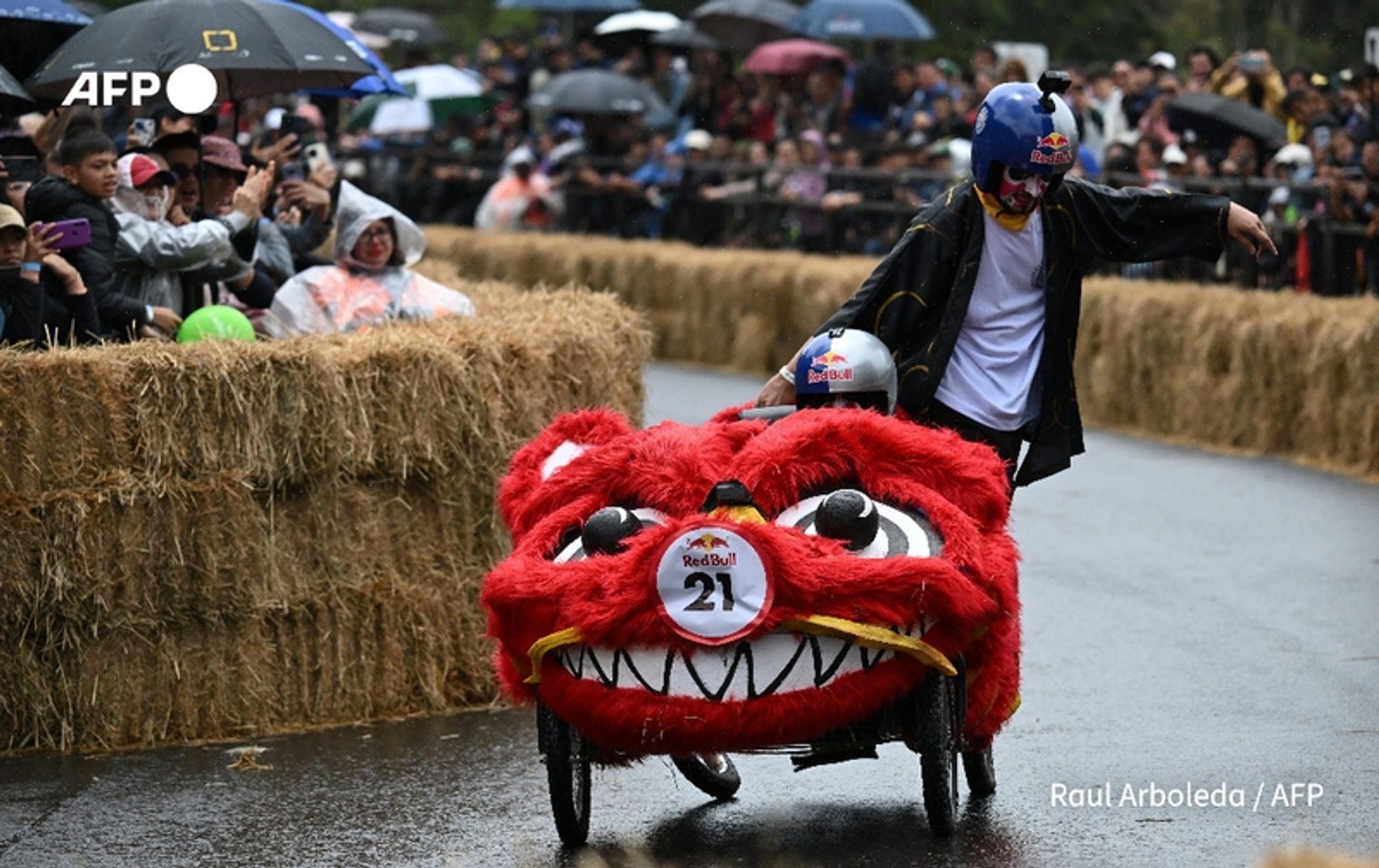 Participants in the annual "Balineras" race in Bogotá, Colombia, navigate a vibrant obstacle course. The foreground features a large, red homemade cart designed to resemble a playful lion. The cart is adorned with exaggerated facial features, including large googly eyes and a wide open mouth, which adds to its whimsical appearance. A racer, wearing a black outfit with a bright red helmet, pushes the cart while pointing enthusiastically towards the crowd. 

Surrounding them is a wet track lined with bales of hay, serving as a safety barrier for spectators. The crowd is visible in the background, many holding umbrellas due to the rainy weather, creating a lively atmosphere despite the drizzle. Some spectators wear casual clothing and appear excited, anticipating the race. The overall setting captures the festive spirit of this unique event, showcasing creativity and community participation in Colombia.