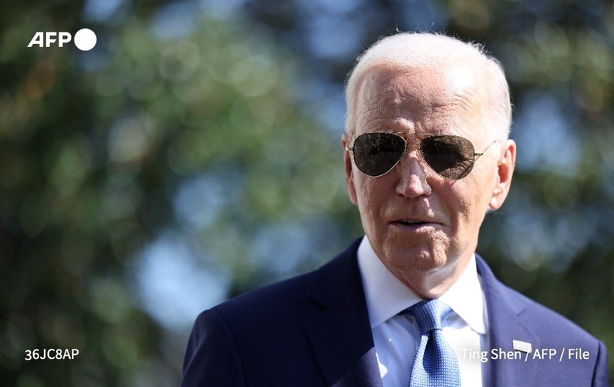 President Joe Biden is depicted in a close-up image, standing outdoors in a bright environment. He is wearing stylish dark sunglasses and a formal attire that includes a navy blue suit and a light blue tie. His white hair is neatly combed, and he has a serious expression as he addresses an unseen audience or camera. The background is blurred, suggesting foliage or trees, indicating a natural outdoor setting. This image captures the context of his remarks about the United States' commitment to Israel's security in the wake of recent events.