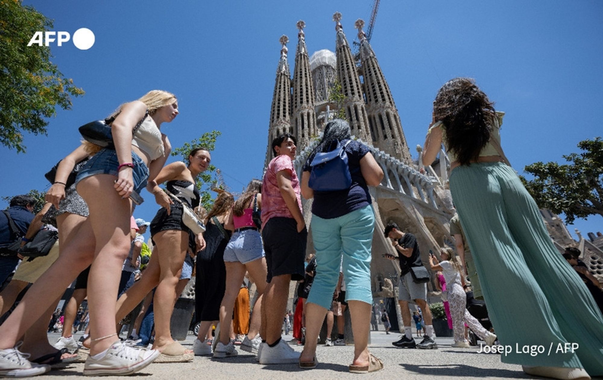 A bustling scene depicting a crowd of tourists gathered in front of the iconic Sagrada Família in Barcelona, Spain. The photograph captures the vibrant energy of summer tourism, with numerous individuals of diverse backgrounds and attire in the foreground. The sunny sky casts bright light on the scene, highlighting the intricate architectural details of the basilica in the background, characterized by its tall spires and unique design. Among the tourists, some are wearing casual summer clothing, such as shorts and tank tops, while others don light dresses or carry backpacks. Nearby trees provide some greenery against the stone structure. The image encapsulates the record influx of visitors as reported in July and August, amidst ongoing protests related to the impact of tourism in the area.