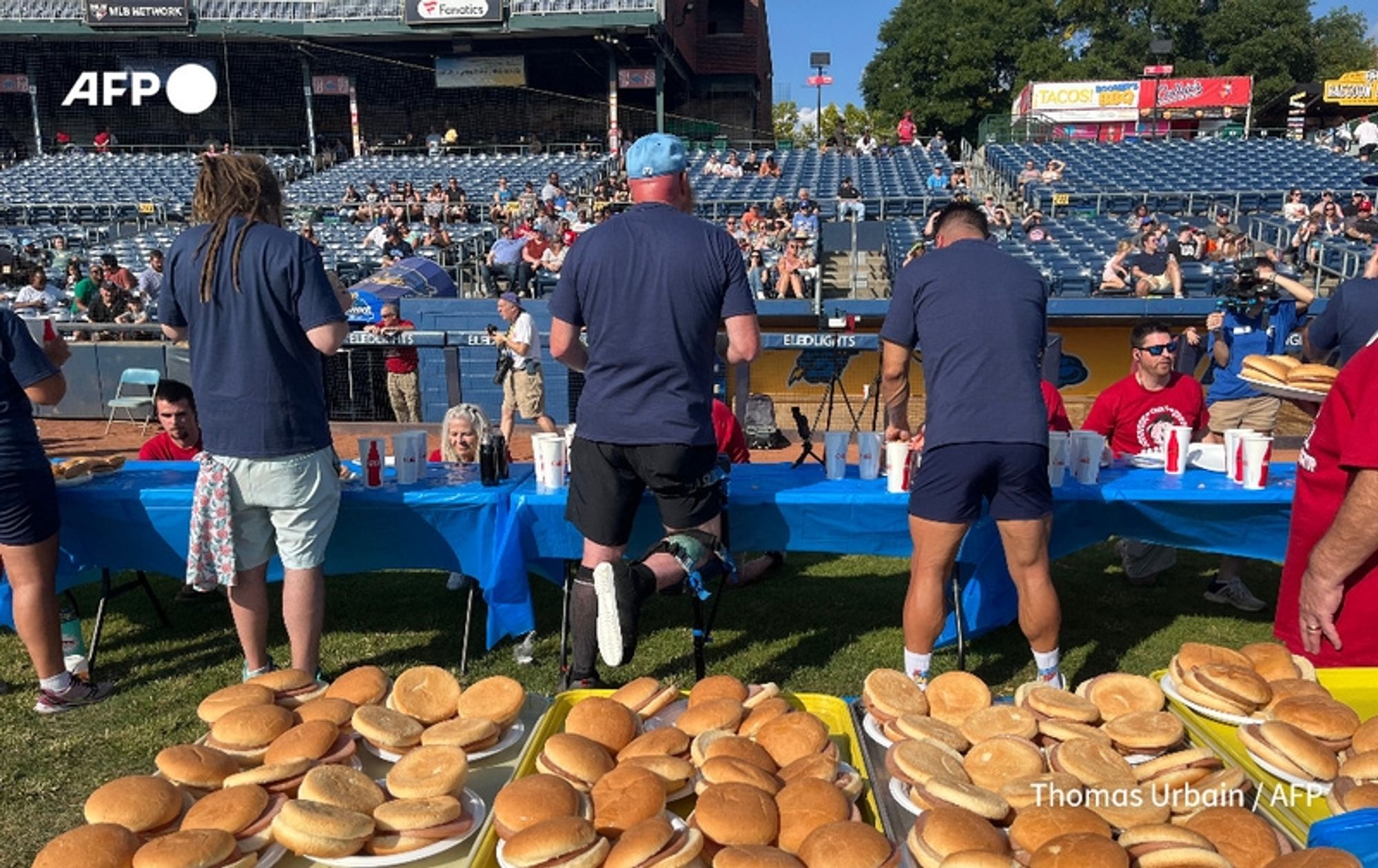 A competitive eating event takes place outdoors, with a large crowd seated in stadium-style seating in the background. In the foreground, several contestants are lined up at a long table draped in blue. Each competitor faces away from the camera, focused on their food. The contestants range in appearance, with one man wearing a black tank top and light shorts, another in a blue shirt and navy shorts, and others in casual attire. A few observers, including a woman with long blond hair and a pink shirt, are seated at the table. The table is laden with stacks of pork rolls, indicating the competitive eating challenge taking place. The setting is lively, with hints of red and blue banners in the background, adding to the festive atmosphere of this unique sport.