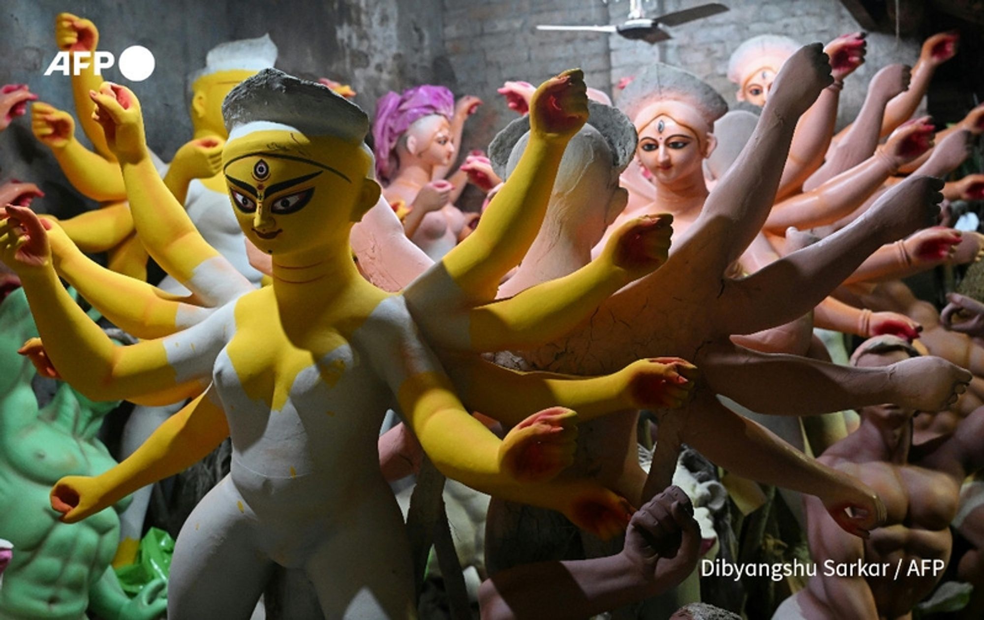 A collection of elaborately crafted clay idols depicting the Hindu goddess Durga in various stages of completion. The central figure of Durga is adorned with multiple arms, each holding different symbolic items, demonstrating her power and divinity. The idols are arranged closely together in a dimly lit workshop, showcasing vibrant colors on some figures, including striking yellows and pinks. The background reveals a rustic wall, enhancing the traditional aspect of the scene, while scattered tools and materials indicate the ongoing process of craft. The overall atmosphere reflects the cultural significance and artistry involved in creating these idols, particularly in the context of upcoming festivities in Kolkata.