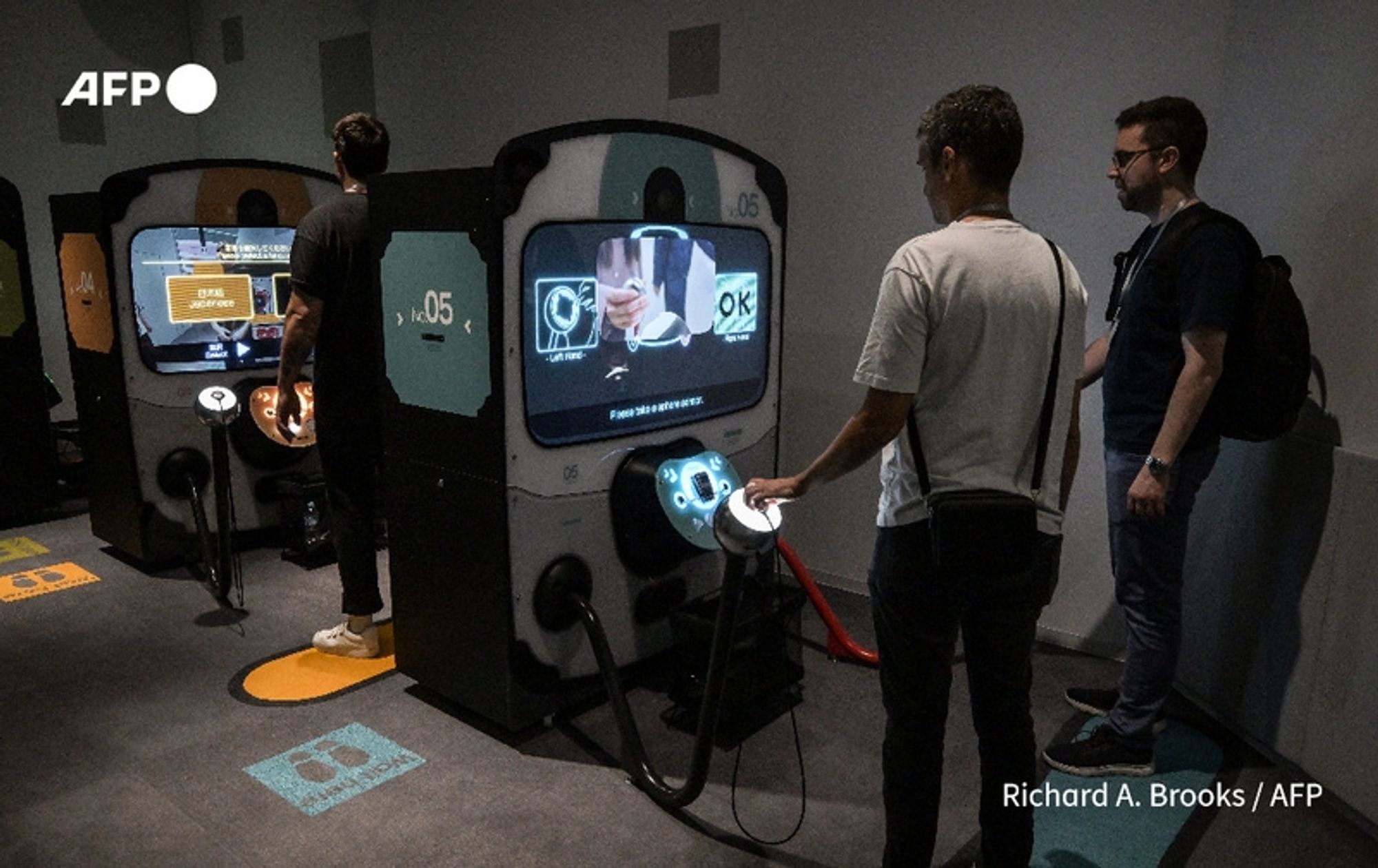 Two individuals are engaged with interactive displays at Nintendo's newly opened museum in Kyoto, which focuses on the company's rich history from its origins in playing cards to its iconic video games. Each display features a large screen with vibrant graphics, showing various gaming interfaces. 

On the left, a person wearing a black shirt and dark pants stands closely, using a joystick attached to one of the consoles, appearing focused on the screen in front of them. The screen shows a colorful interface related to gaming. 

To the right, another individual with short hair, wearing a light gray t-shirt and dark pants, examines a different display. A third person, clad in a dark shirt and holding a bag, observes the interaction with interest. The floor is decorated with a modern design, including Nintendo's iconic logo, adding to the museum's engaging atmosphere. Soft lighting enhances the exhibits, creating an inviting space for visitors to explore Nintendo's legacy.