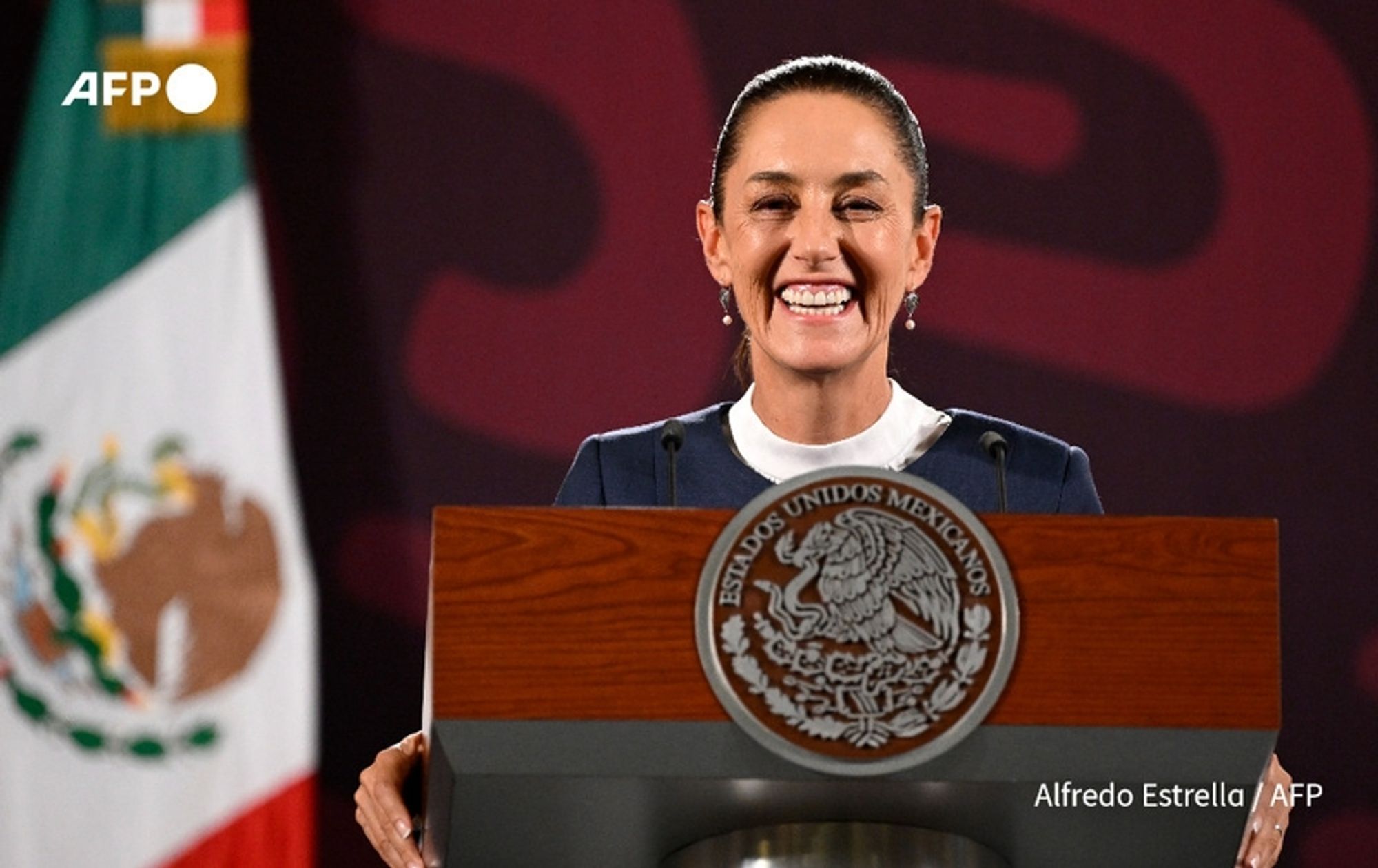 A woman stands at a podium with a large emblem featuring the Mexican coat of arms, which includes an eagle perched on a cactus. She has long, dark hair pulled back and is wearing a dark suit with a white shirt. The woman is smiling broadly, conveying a sense of joy and accomplishment. Behind her, there is a Mexican flag with green, white, and red stripes, along with a coat of arms on the left side of the image. The background consists of a bold, abstract design in shades of red and dark hues. The setting appears to be an official event, possibly a swearing-in ceremony. The overall atmosphere is celebratory and significant, highlighting her historic role as the first female president of Mexico.