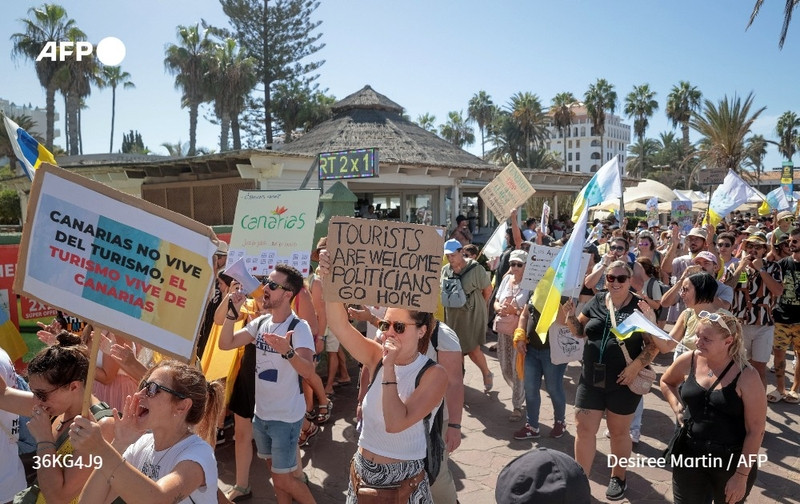 A large crowd of demonstrators is gathered on a sunny day in a location featuring palm trees and a tropical atmosphere, typical of the Canary Islands. The scene shows a diverse group of people, some wearing sunglasses and casual summer clothing, actively participating in a protest against mass tourism. Various colorful banners and signs are being held high by the protesters, including one that reads "CANARIAS NO VIVE DEL TURISMO; EL TURISMO VIVE DE CANARIAS" and another that states "TOURISTS ARE WELCOME POLITICIANS GO HOME." The background reveals structures with thatched roofs and greenery, indicative of a lively tourist destination. The enthusiasm of the demonstrators is apparent as they wave flags and shout, showcasing their strong sentiments regarding tourism in their homeland. The atmosphere conveys a sense of solidarity among the participants in their call for change.