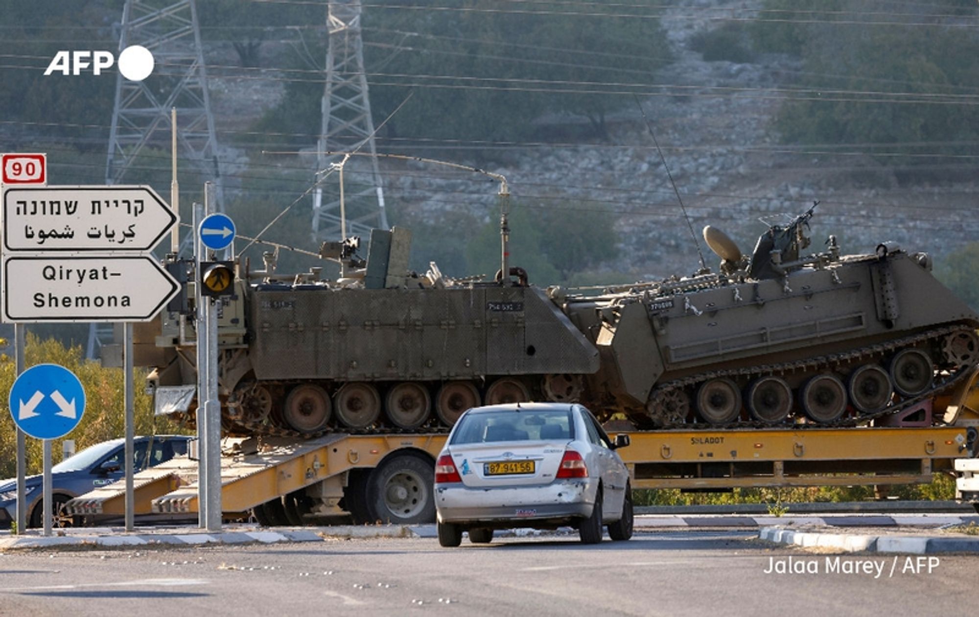 A military vehicle, resembling an armored personnel carrier, is seen on a flatbed truck at a junction. The setting is a road sign indicating directions to Qiryat Shemona, with both Hebrew and Arabic text displayed. A silver car, parked nearby, appears to be in motion, positioned in front of the truck. In the background, a hilly landscape with sparse vegetation can be observed, along with electricity pylons standing tall. The scene conveys a tense atmosphere amidst ongoing military operations, highlighting the military presence and logistics related to the conflict in Lebanon involving Hezbollah.