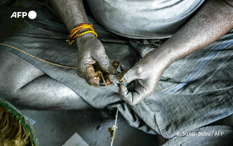 A close-up image of a worker's hands engaged in the manual production of fireworks in an Indian fireworks factory. The hands are heavily soiled, glistening with a layer of fine dust, indicative of the materials used in fireworks manufacturing. The worker is wrapping thin lengths of silver fuse and using a sticky mixture to adhere it, showcasing dexterous hand movements. The hands are adorned with yellow and red thread bangles, adding a cultural touch. The worker is seated on the floor, their gray clothing visible in the background, blending into the dimly lit factory setting that emphasizes the labor-intensive nature of preparing for the Diwali festival. Various materials and components of fireworks are scattered around, enhancing the sense of a busy workspace.