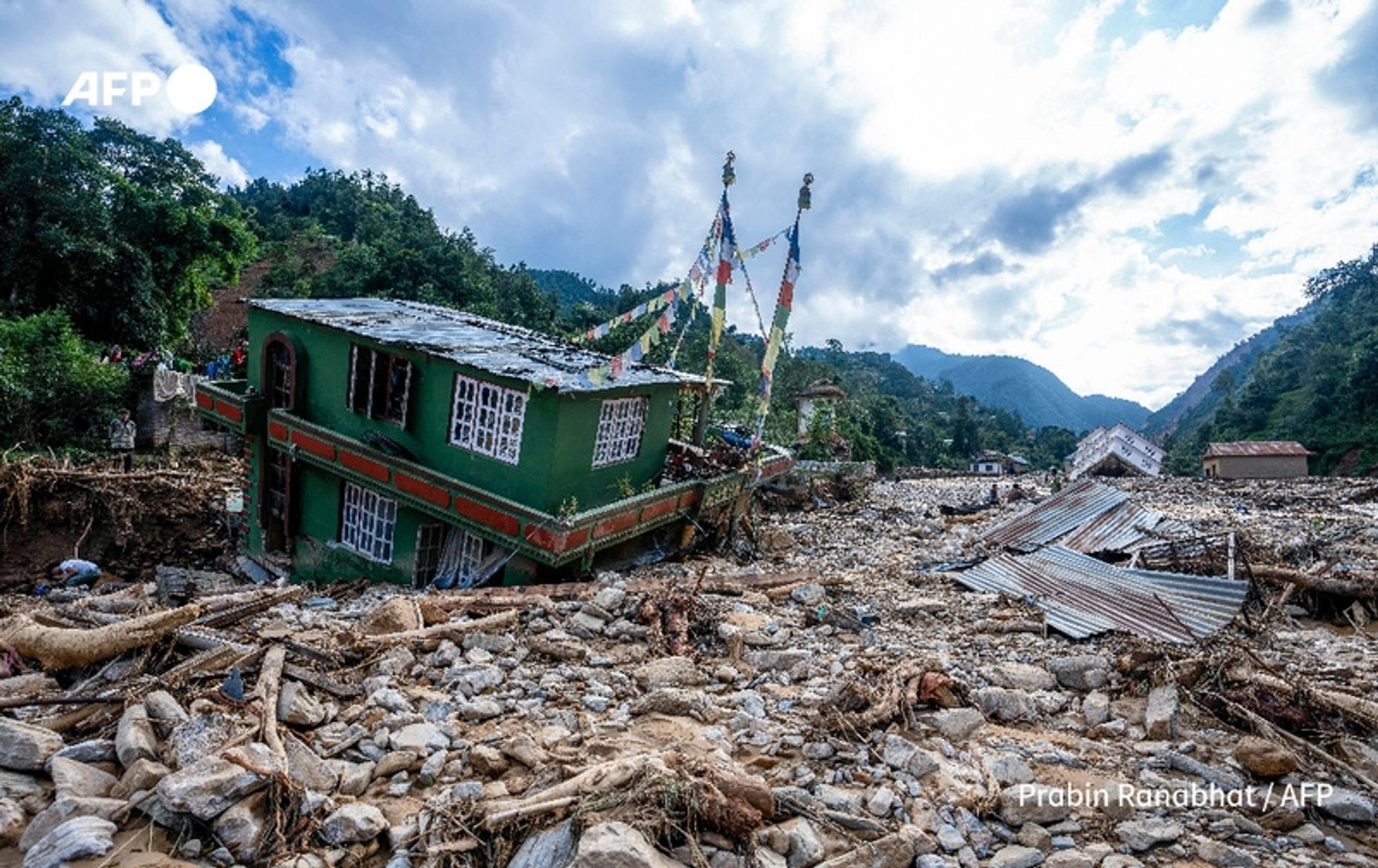 A scene depicting the aftermath of monsoon floods in Nepal, showcasing a densely debris-strewn landscape. In the foreground, the remains of a partially collapsed green building with white windows is visible, leaning precariously to one side. The building features colorful prayer flags strung from its roof, fluttering gently in the breeze. Surrounding the structure, large stones, mud, and fallen logs create a chaotic scene, indicating the force of the floodwaters that have receded. In the background, several other damaged homes can be seen, along with a mountainous landscape under a blue sky with scattered clouds. The image conveys the destruction wrought by the floods, with search and rescue teams likely engaged in recovery efforts among the debris.