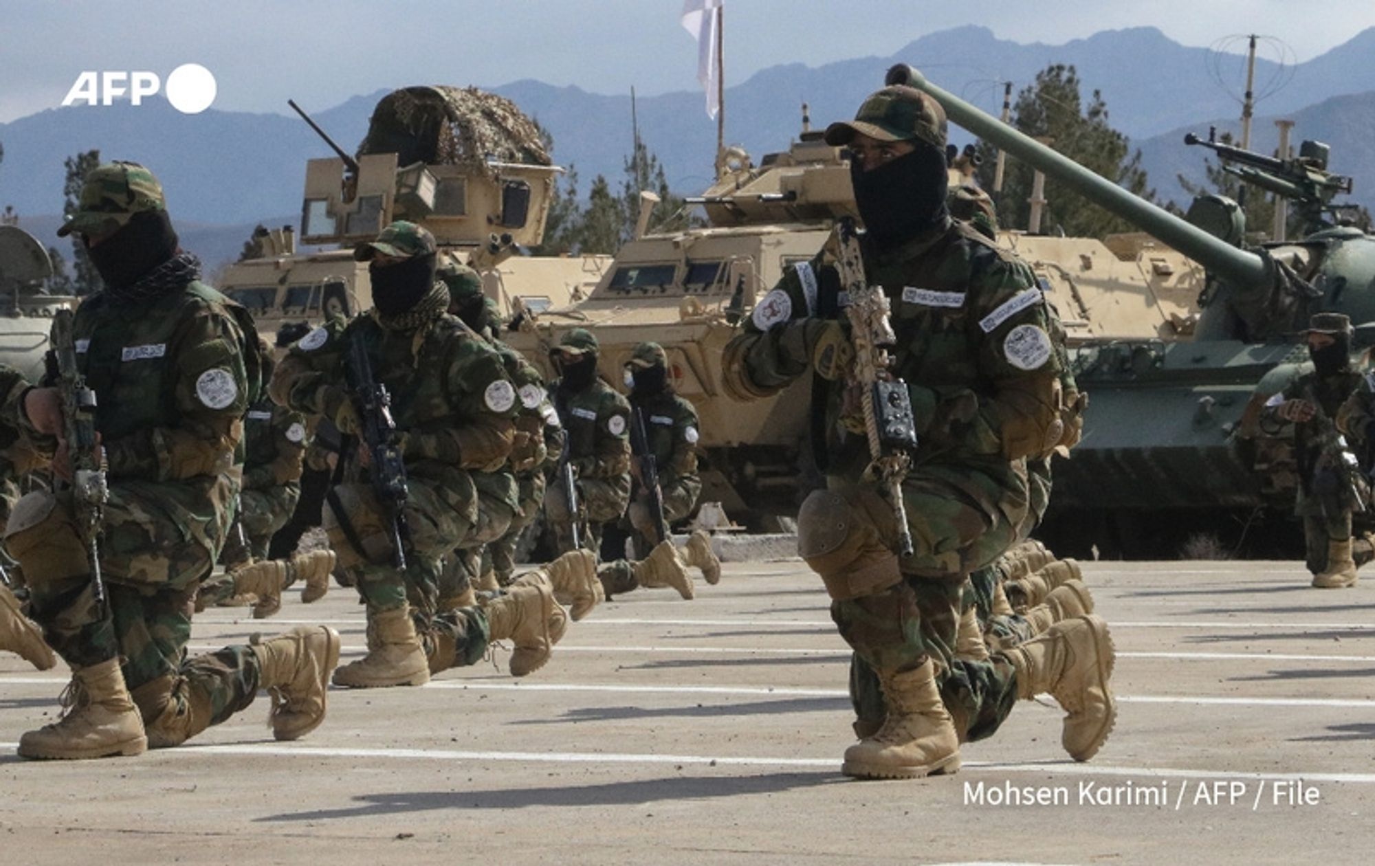 A group of soldiers wearing military camouflage uniforms and black masks marches in a formation. They are holding weapons and appear to be in a training exercise or official demonstration. The background features military vehicles, including armored personnel carriers and tanks, set against a backdrop of mountains under a cloudy sky. The atmosphere conveys a sense of readiness and discipline. The soldiers are positioned on a paved surface, and there are flags and trees visible in the distance. The image reflects the context of Afghanistan's security situation, highlighting the Taliban's military presence in response to threats posed by the Islamic State group.