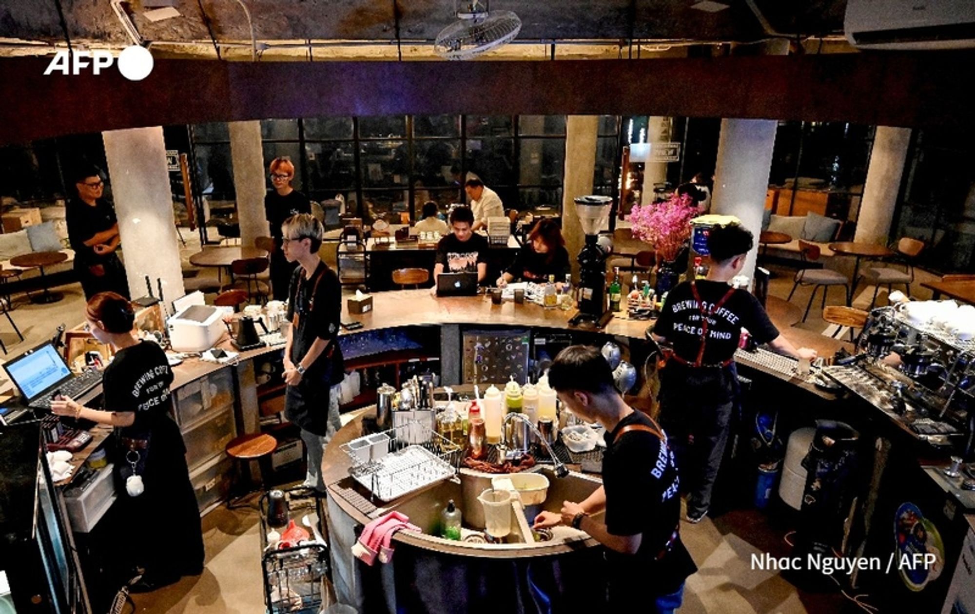 A vibrant café scene featuring several young Vietnamese entrepreneurs engaged in various activities. The image showcases a central, circular bar-like counter with a coffee preparation area and multiple workstations. In the foreground, two individuals collaborate closely, one at a sink washing equipment while the other prepares drinks. Behind them, more young adults work on laptops, deeply focused on their tasks. The café has a modern, industrial aesthetic, with bare concrete walls and wooden furniture. A large window lets in natural light, illuminating the workspace. Various coffee-making tools and ingredients are scattered across the counter, indicating a busy but organized environment. The atmosphere appears energetic and collaborative, highlighting a new wave of entrepreneurship in the coffee industry among the younger generation.