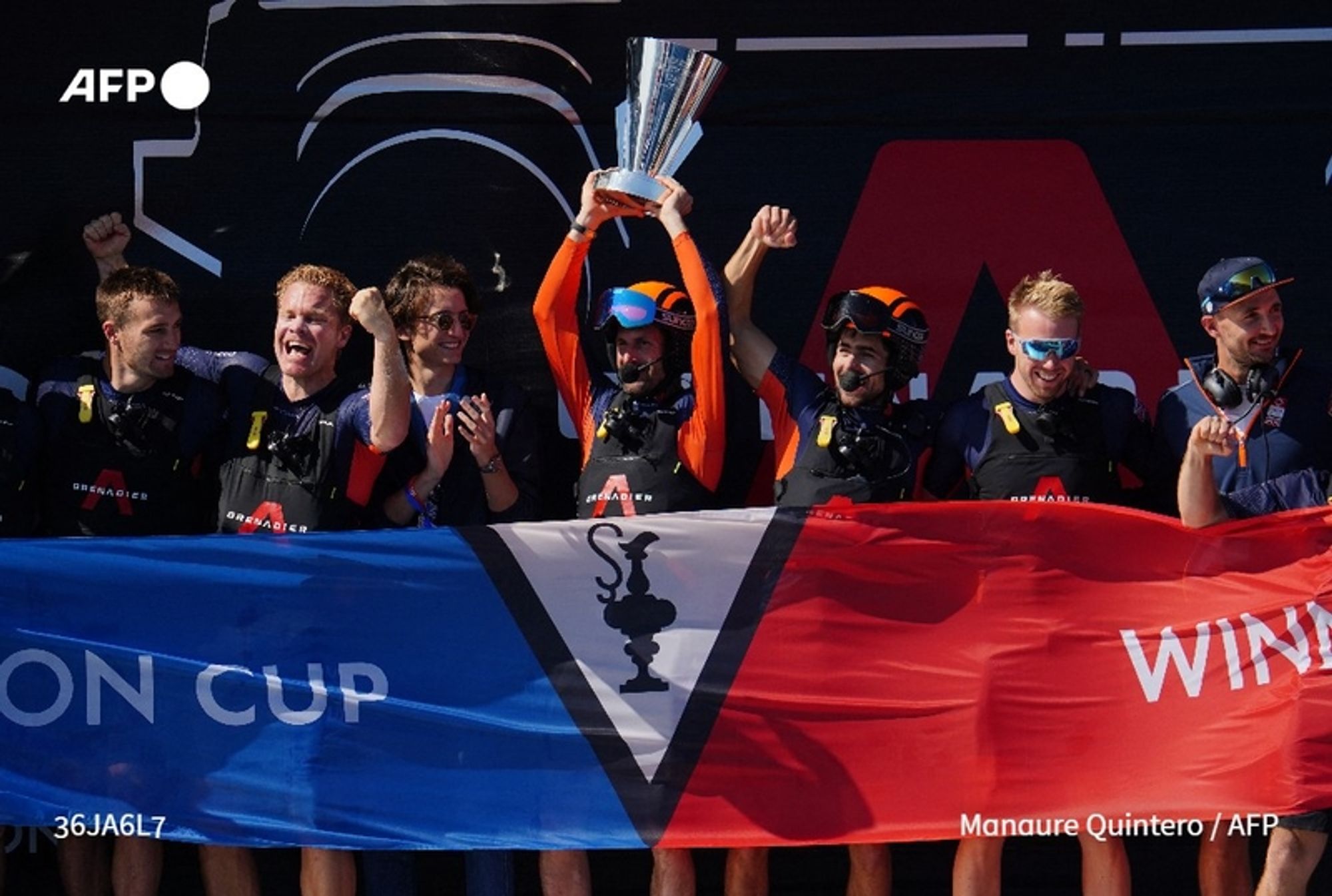 A celebration scene featuring a group of six jubilant sailors, all wearing dark sailing gear with life jackets, standing together on a podium. The central figure, raising a trophy high above their head, is wearing an orange cap and an orange top. They are flanked by teammates who are expressing excitement with raised arms and cheering. One sailor holds a large flag, predominantly blue and red, that features a diagonal white section with a black emblem. The backdrop includes a large logo, possibly related to the event. The atmosphere is festive, capturing a moment of victory, following Britain's qualification for the America's Cup after defeating Italy in the Louis Vuitton Cup off the coast of Barcelona.