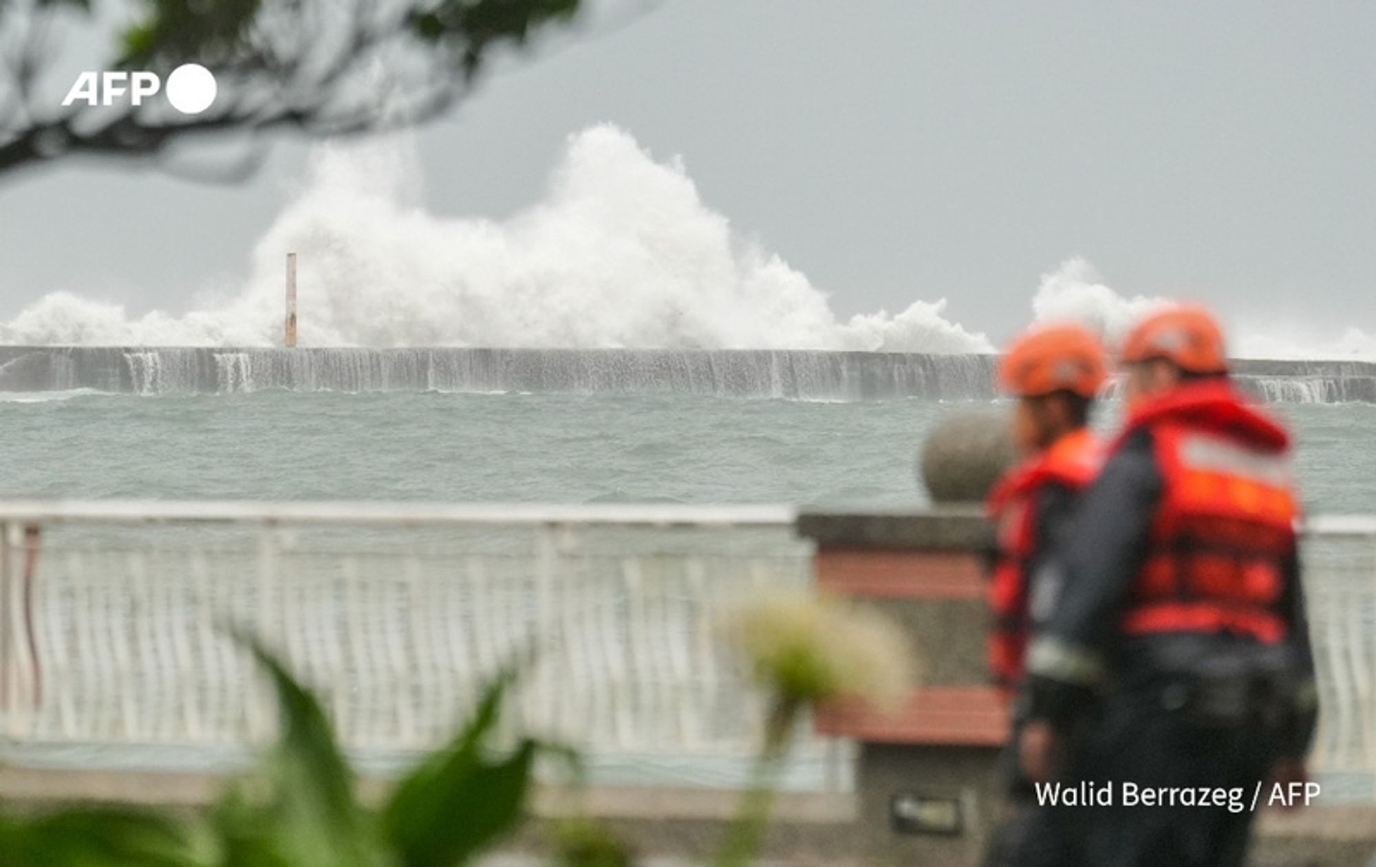 Two rescue workers in bright orange helmets and vests are seen walking along a coastal promenade. The ocean is visibly turbulent, with large waves crashing against a breakwater in the background, sending up sprays of water. The sky is overcast and gray, indicating adverse weather conditions. In the foreground, part of a railing and some greenery can be seen, partially obscuring the view of the workers. The scene conveys a sense of urgency and the impact of Typhoon Krathon on the island, highlighting the danger as the storm approaches.