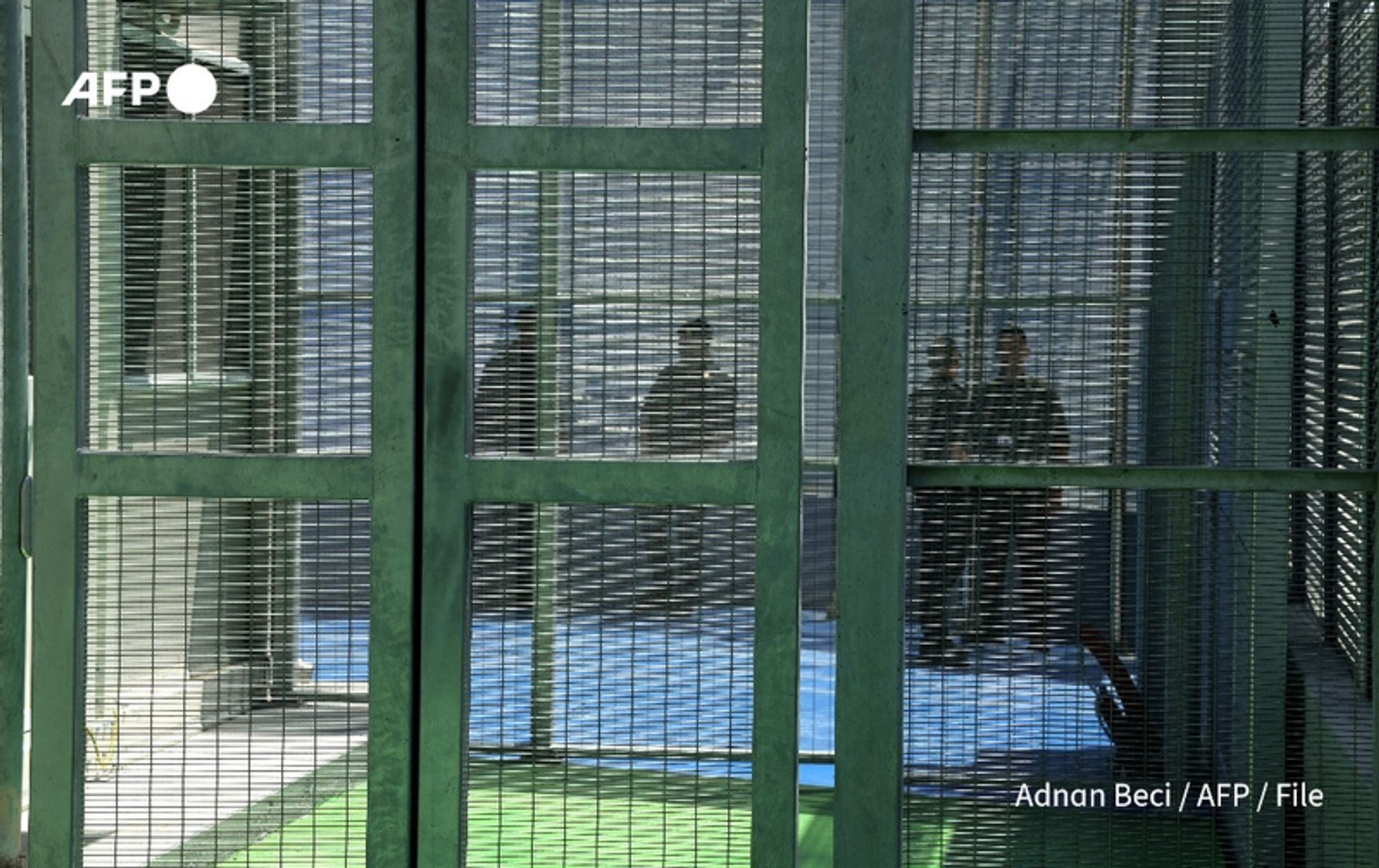 A fenced area with green metal gates and mesh panels, creating a sense of confinement. Inside the enclosure, silhouettes of several individuals can be seen, suggesting they are standing or walking. The background is partially visible, featuring blue flooring and a bright, open space beyond the fencing. The overall tone of the image conveys a somber and restrictive environment, highlighting themes of migration and enforcement. The photo appears to emphasize current discussions surrounding deportation and immigration policies in Europe, particularly in light of recent political shifts. The image is credited to Adnan Beci / AFP / File.