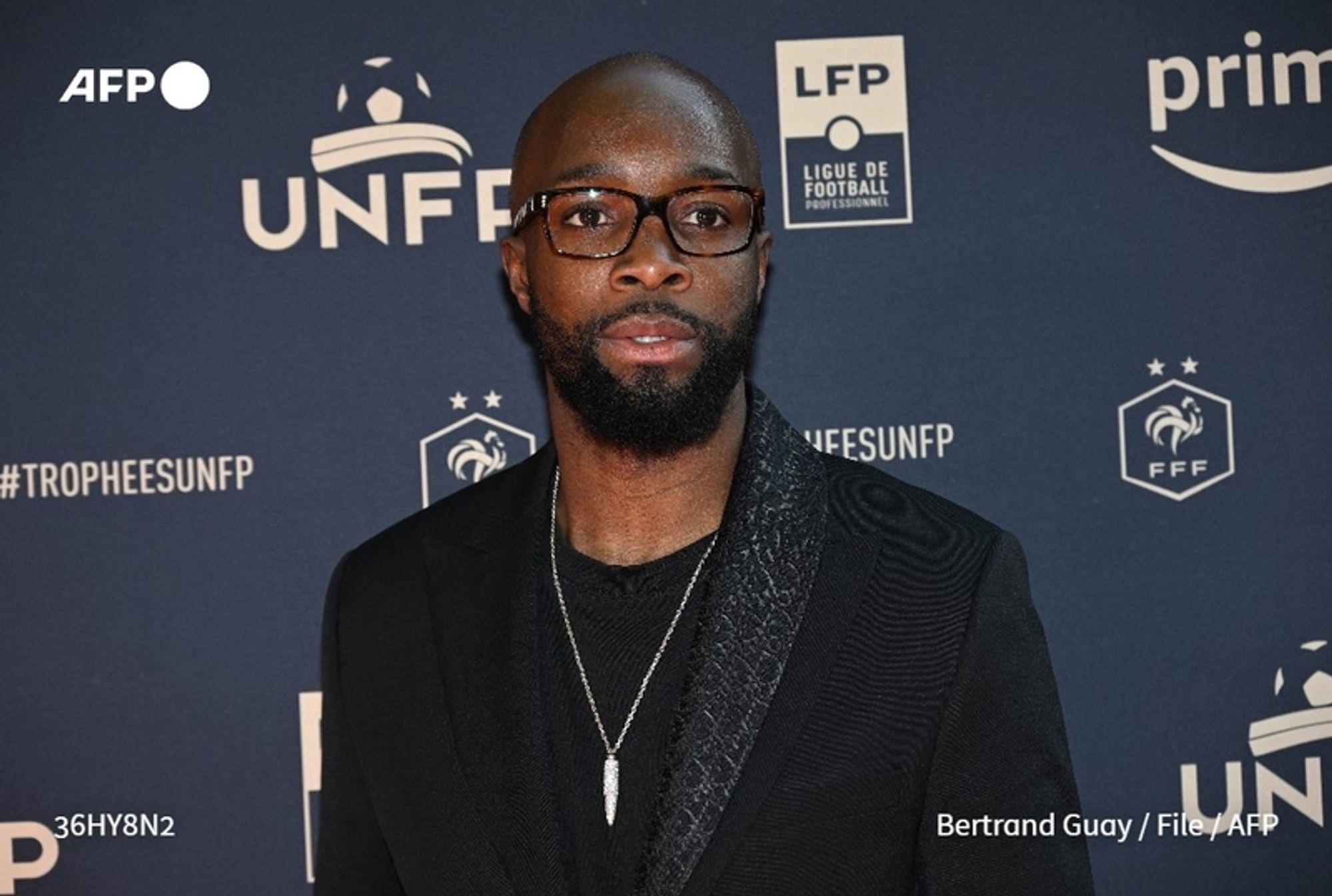 A well-dressed man with a short beard and glasses stands in front of a dark blue backdrop featuring various logos, including the UNFP (Union Nationale des Footballeurs Professionnels) and the FFF (Fédération Française de Football). He is wearing a black jacket with a textured lapel over a black shirt. The man's expression is serious, and he is looking directly at the camera. His hair is closely cropped, and he has a necklace visible around his neck. The image conveys a formal event atmosphere, likely related to football, given the presence of logos associated with the sport.