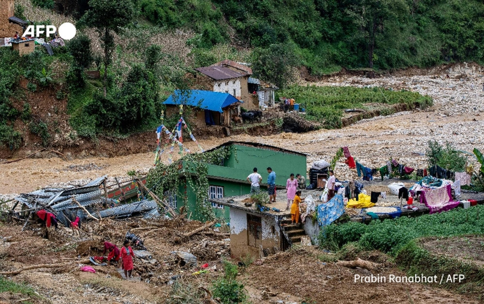 A scene depicting the aftermath of monsoon floods in Nepal. The image shows a landscape of damaged homes and debris scattered across the area. In the foreground, a group of rescue workers and local residents are sifting through the wreckage, which includes twisted metal, wooden beams, and remnants of structures. Some people are seen wearing bright clothing, indicating their involvement in the cleanup efforts. 

In the background, partially destroyed houses can be seen, with roofs missing and sections of walls collapsed. A blue-tarp covered structure is visible among the rubble, alongside several colorful prayer flags fluttering in the breeze, suggesting the region's cultural significance. The ground is covered with mud and debris, with patches of greenery pushing through, hinting at the resilience of nature. The overall mood conveys a sense of urgency and determination as the community comes together to recover from the devastating floods that have impacted the region.