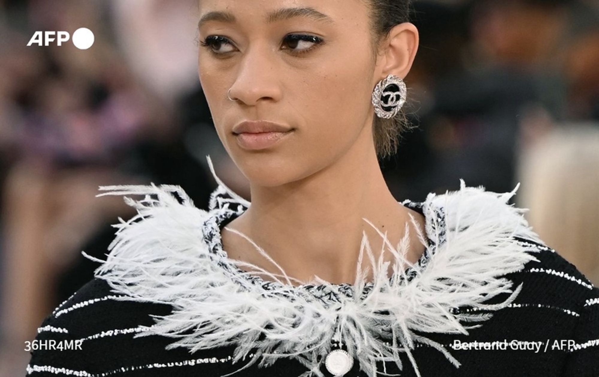 A close-up of a model with a serious expression, showcasing her elegance at a fashion show. She has light brown skin and wears her dark hair in a neat, sleek style. The model is dressed in a black and white striped coat with a dramatic feathered collar that adds a touch of luxury and playfulness to her look. Her outfit features embroidered details, enhancing the sophisticated design. She accessorizes with a large, shiny Chanel logo earring that stands out against her skin. The background is slightly blurred, indicating a bustling fashion event atmosphere, consistent with the grandeur of the Grand Palais during Paris Fashion Week.