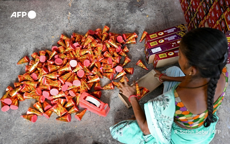 A woman sits on the floor in a factory, surrounded by a large pile of cone-shaped sparklers wrapped in vibrant red paper. The sparklers, which have circular bases in pink, are arranged haphazardly around her. She is focused on sorting or packing them, using a cardboard box nearby. The woman has dark hair styled in a braid and wears a light teal sari adorned with colorful patterns on the sleeves. In the background, there are stacks of boxes printed with designs, indicating the festive theme associated with the upcoming Diwali celebration. The setting has a concrete floor, typical of a working environment, and there is a bright, industrious atmosphere as preparations for the Hindu festival of lights are underway.