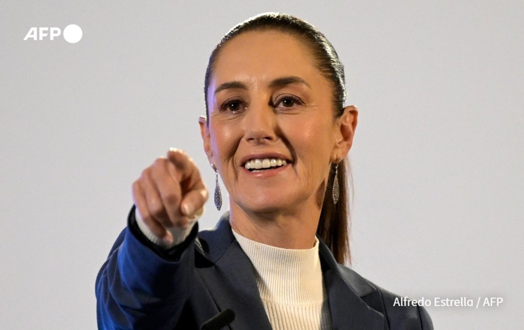 Claudia Sheinbaum, the President of Mexico, is depicted in a close-up shot, enthusiastically gesturing while speaking. She has long brown hair slicked back and is wearing a dark blue blazer over a light-colored turtleneck sweater. Her expression is confident and engaged as she points towards the audience with her right hand, smiling broadly. Sheinbaum's earrings are visible, adding a touch of elegance to her professional attire. The background appears neutral and slightly blurred, emphasizing her presence in the image. The photograph suggests an atmosphere of leadership and determination, coinciding with her recent announcement of proposals aimed at enhancing gender equality in Mexico.