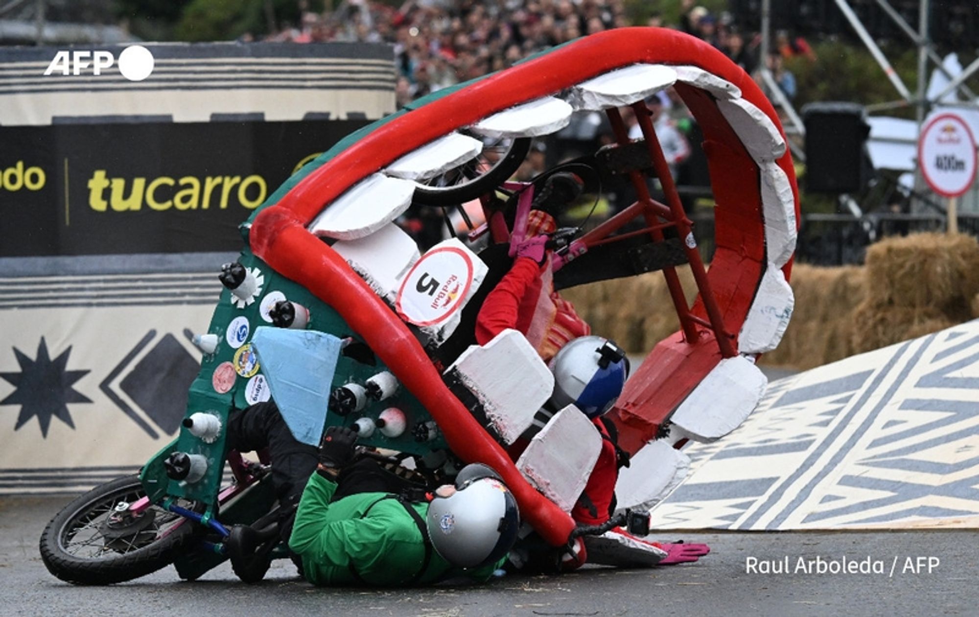 Participants in the annual "Balineras" race in Bogota, Colombia, are seen engaging in a vibrant and energetic scene. The image captures several racers riding homemade carts, one of which is dramatically overturned. The cart, decorated in bright colors with a red top and various stickers, displays a playful design that resembles a large mouth. Two participants are sprawled on the ground, wearing helmets and bright clothing, indicating the thrilling and chaotic nature of the race. The background features a cheering crowd and a festive atmosphere, contributing to the lively spirit of this unique obstacle course event. The racecourse is marked with distinctive patterns and signage, enhancing the colorful and adventurous environment of the competition.