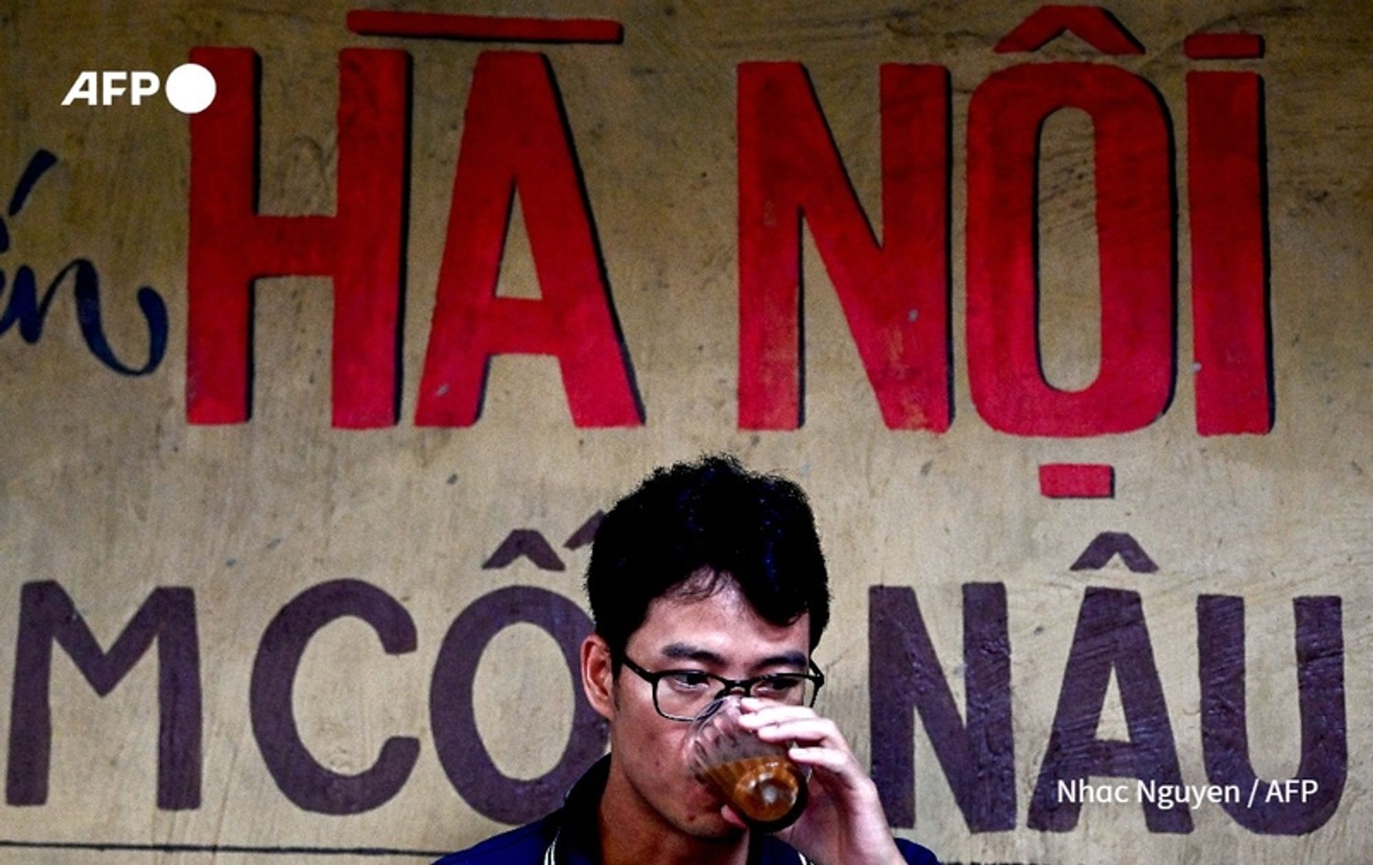 A young Vietnamese man is sitting in front of a weathered wall painted in earthy tones. Prominently displayed behind him is bold red text that reads "HÀ NỘI." He is wearing glasses and has dark, slightly tousled hair. As he drinks from a glass filled with a brown beverage, his expression is contemplative. The man's casual attire contrasts with the traditional expectations depicted by the surroundings. Various designs and characters in the wall's paint hint at a local coffee culture, reflecting the entrepreneurial spirit amidst generational family expectations regarding work. The scene captures the essence of young Vietnamese entrepreneurs embracing coffee-making as a means of challenging societal norms.