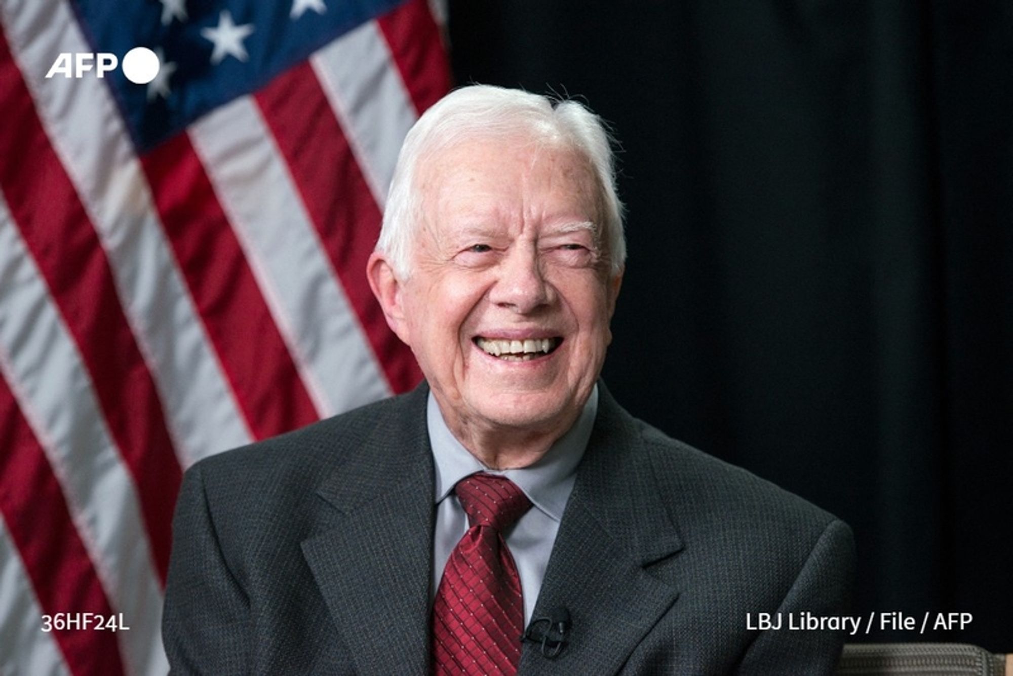 Jimmy Carter, the 39th President of the United States, is seated and smiling in a warm and inviting manner, celebrating a significant milestone—his 100th birthday. He has light gray hair and wears a dark suit jacket over a collared shirt, complemented by a red tie. Behind him, the American flag proudly displays its stars and stripes, providing a patriotic backdrop. The setting appears formal, with a dark background that highlights his approachable expression and the joyful occasion. The overall atmosphere conveys a sense of celebration and honor for his extraordinary life and contributions.