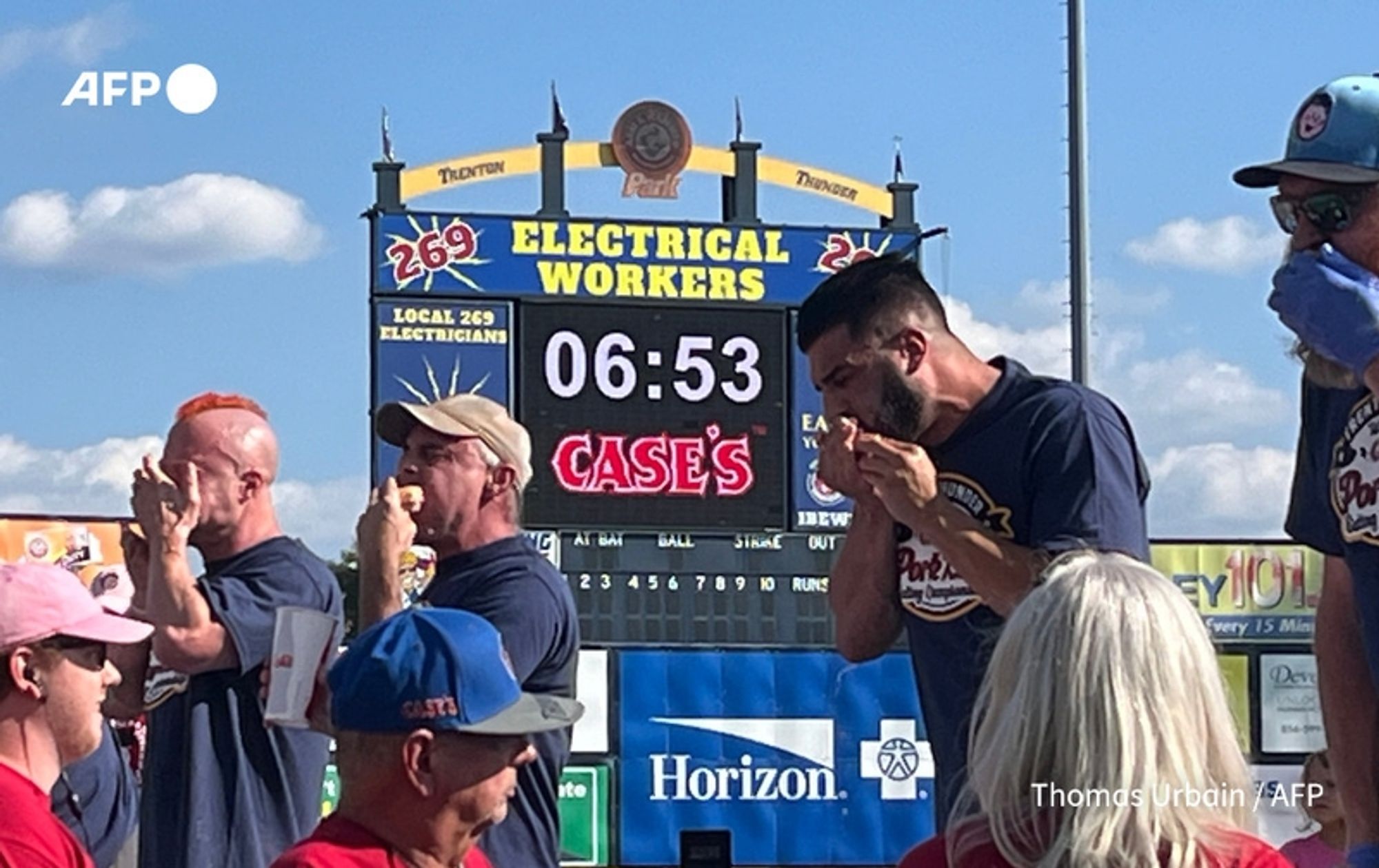 A vibrant outdoor scene at a competitive eating event, showcasing several male contestants engaged in the challenge of consuming pork rolls. In the foreground, one contestant in a dark shirt appears to be preparing to eat, with his hand held up to his mouth, while a second contestant beside him has a focused expression, indicating concentration. They are surrounded by spectators, some wearing event-themed clothing. In the background, a large digital scoreboard displays a timer showing 6 minutes and 53 seconds remaining, with the words "ELECTRICAL WORKERS" prominently featured above it. The event takes place under a clear blue sky, creating a lively and festive atmosphere.