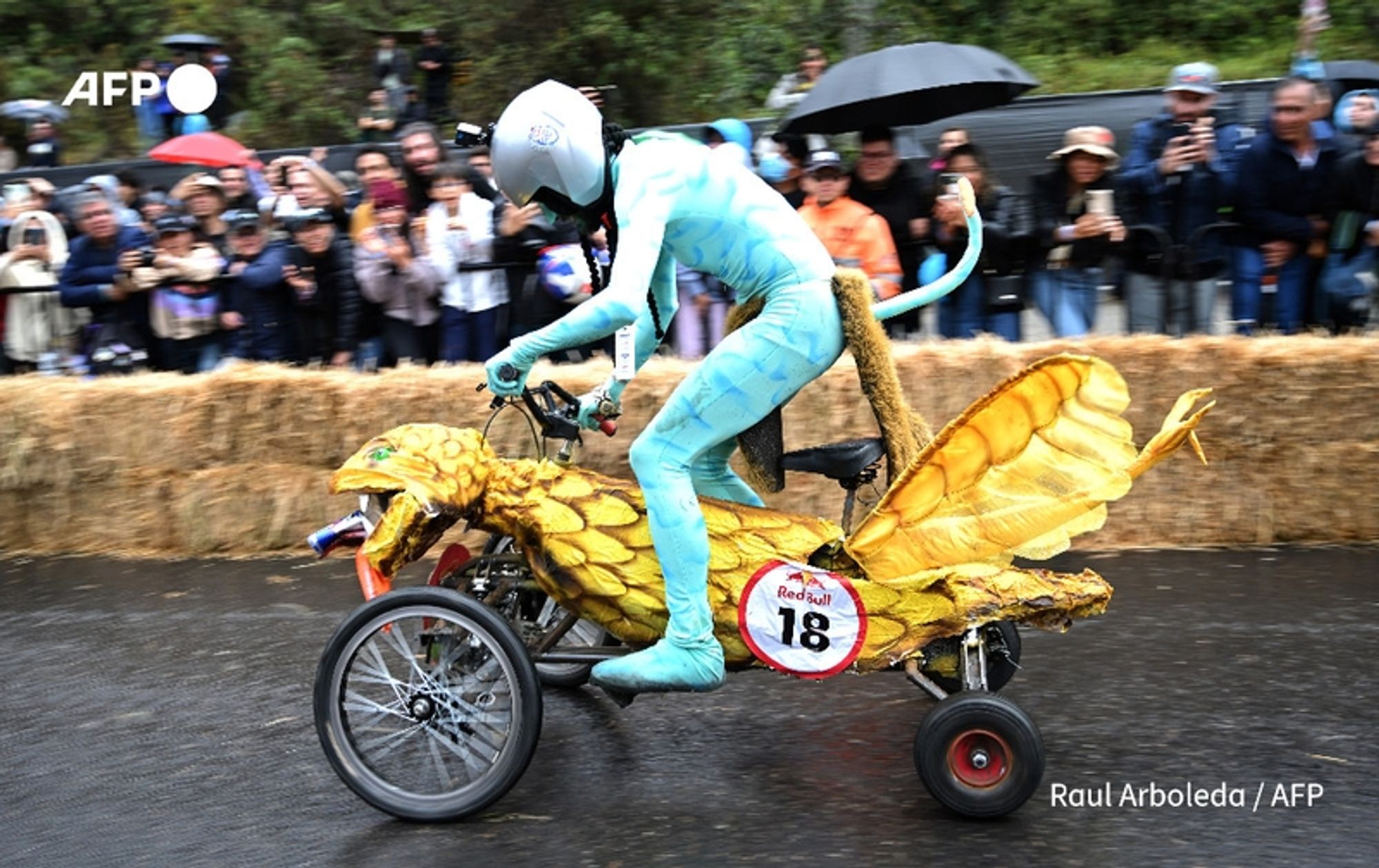 A participant is racing in a homemade cart during the annual "Balineras" race in Bogotá, Colombia. The cart is creatively designed to resemble a fantastical creature, featuring large, vibrant yellow wings resembling a dragonfly or mythical beast. The racer, dressed in a bright blue bodysuit that matches the color scheme of the cart, is wearing a white helmet for safety. They are positioned mid-race, leaning forward as they maneuver the cart along a wet road. In the background, a crowd of onlookers dressed in various outfits watches intently, some holding umbrellas, indicating light rain during the event. Bales of hay line the side of the track, and the atmosphere is festive and energetic as participants engage in this whimsical obstacle course. A circular red and white number plate marked "18" is affixed to the cart, showcasing the participant's number.