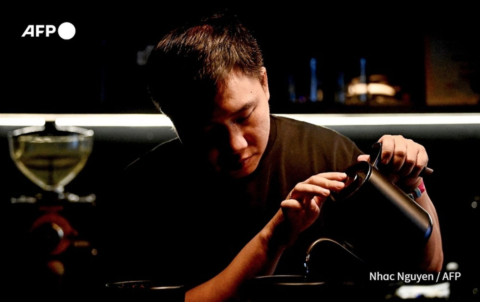 A young Vietnamese entrepreneur focuses intently on pouring coffee from a sleek, black kettle into a cup. The setting is dimly lit, with subtle, warm lighting enhancing the ambiance of the coffee shop. Various coffee-making equipment is visible in the background, including a glass brewing apparatus and neatly arranged utensils. The entrepreneur's serious expression conveys dedication and passion for their craft, as they engage in the meticulous process of preparing coffee. Their understated black shirt contrasts with the glow from the brewing equipment, emphasizing their concentration on the task at hand. This scene represents a new generation of young Vietnamese individuals challenging traditional workplace expectations by embracing coffee culture as a means of entrepreneurship.