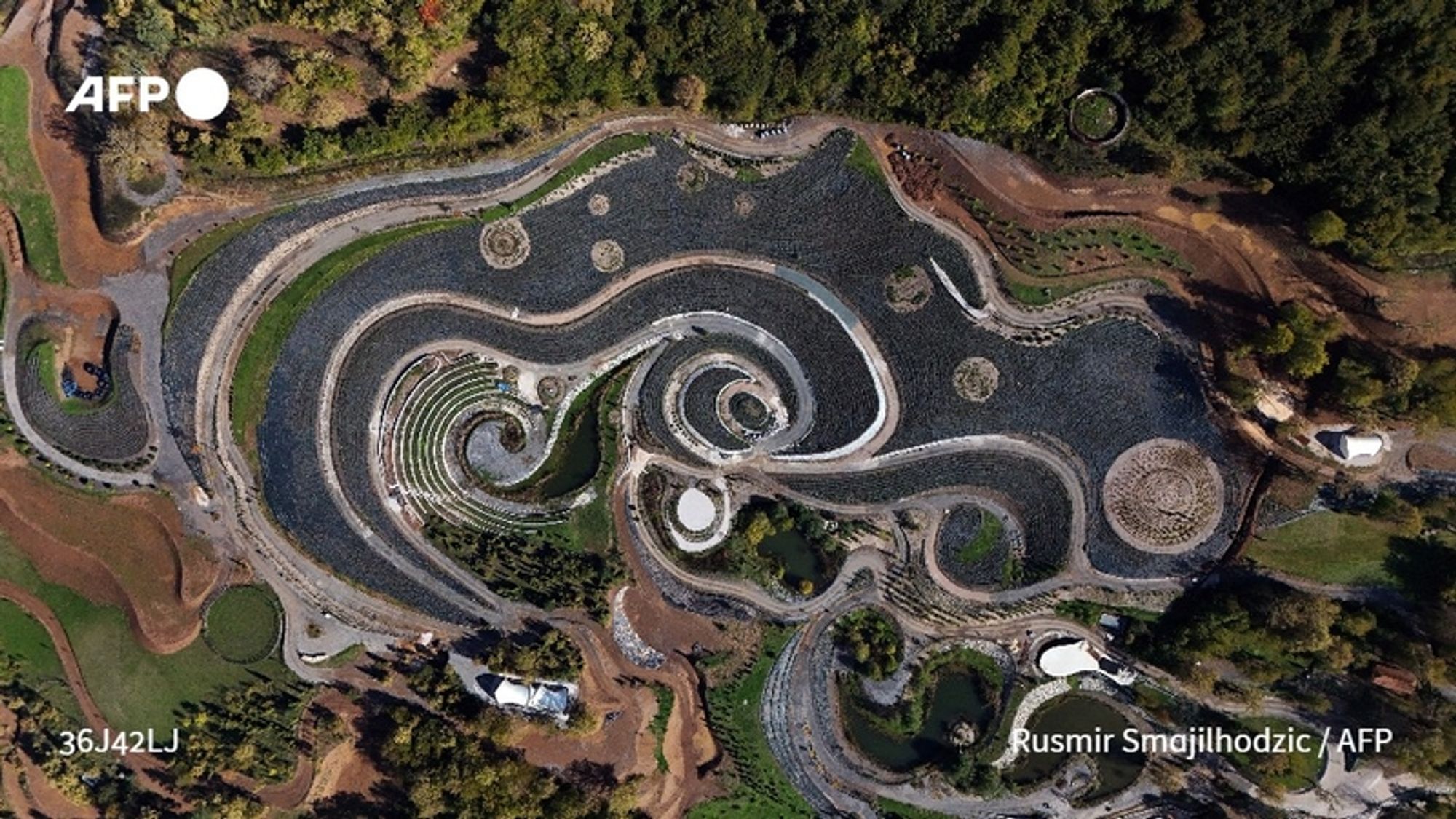 Aerial view of a large, beautifully landscaped area resembling Vincent Van Gogh's "Starry Night". The intricate design features swirling patterns outlined in different colors and textures of plants, mimicking the iconic brush strokes of the painting. Various plant beds create the illusion of movement, with clusters of vibrant greens contrasting against darker soil. Small ponds are interspersed throughout, reflecting the surrounding landscape. The garden's layout includes circular and spiral shapes that evoke the stars and sky depicted in the original artwork, enhancing the connection to Van Gogh's masterpiece. Surrounding the garden is a natural landscape of trees and plants, contributing to the serene and artistic atmosphere of this living homage to art.