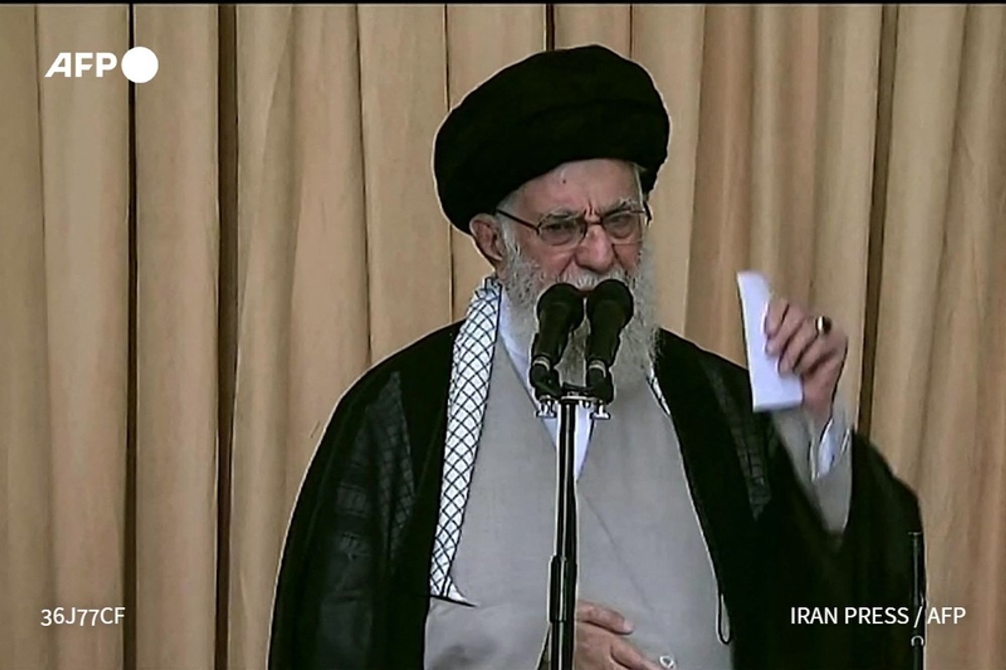 A middle-aged man with a long beard, wearing a black turban and traditional clerical attire, stands at a podium with two microphones in front of him. He appears to be giving a speech, holding a piece of paper in his right hand. The background features light-colored curtains, suggesting a formal setting. The man looks serious and engaged, as he emphasizes his points during the address. He has glasses and a solemn expression, underscoring the significance of his message. The image conveys a sense of authority and determination.