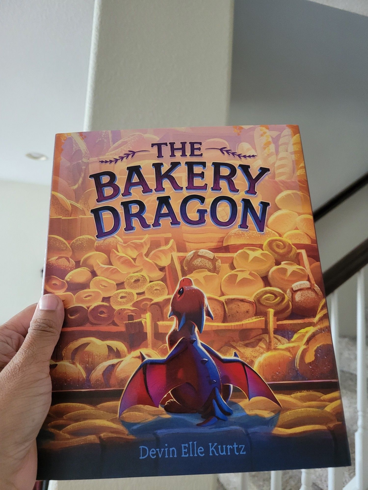 A book with a small red dragon looking into a bakery titled The Bakery Dragon by Devin Ellie Kurtz.