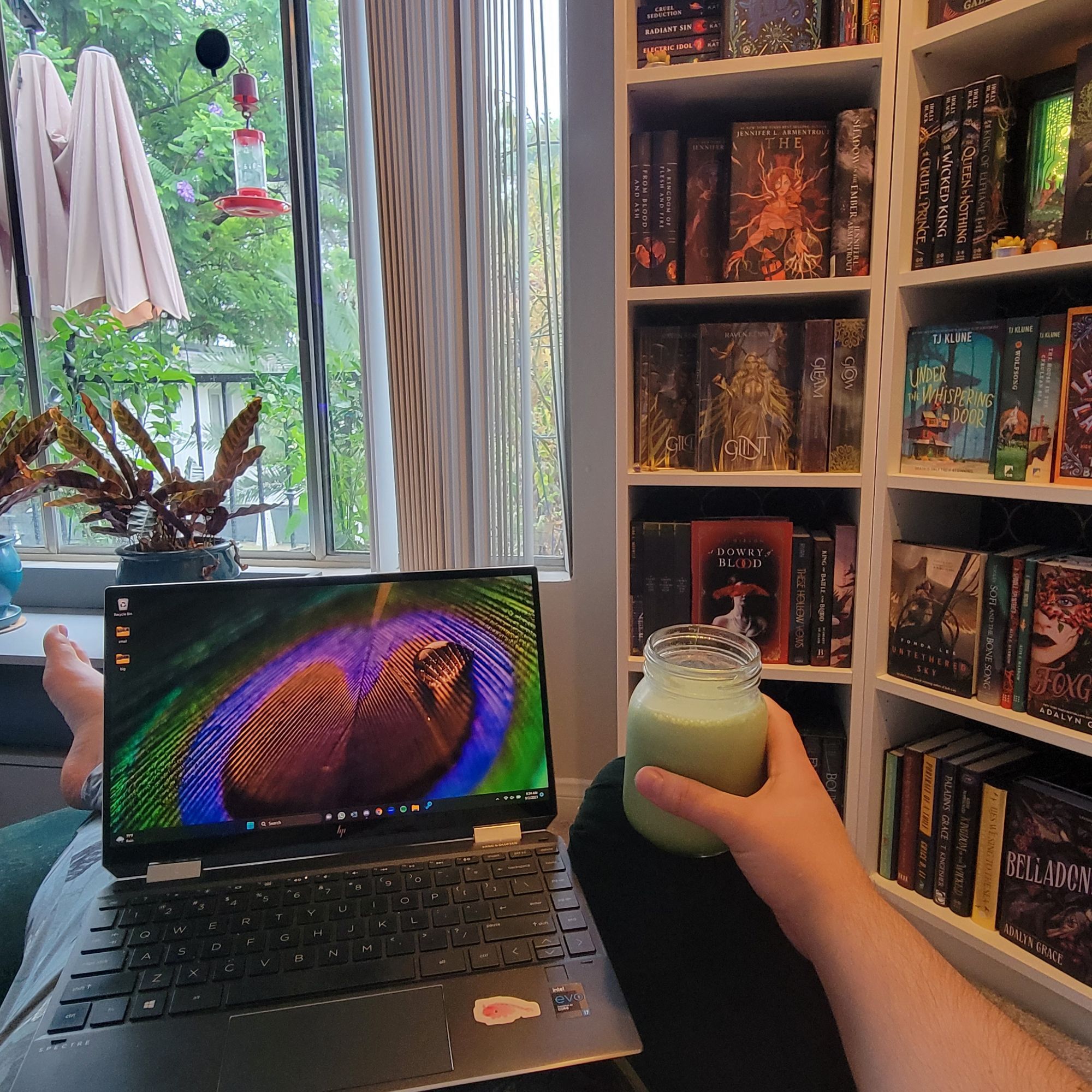 A black laptop sitting on a lap, in a green armchair, an arm holding a glass jaw of matcha on the armrest. In the background are bookshelves to the right, a window looking out on rain and trees to the left.