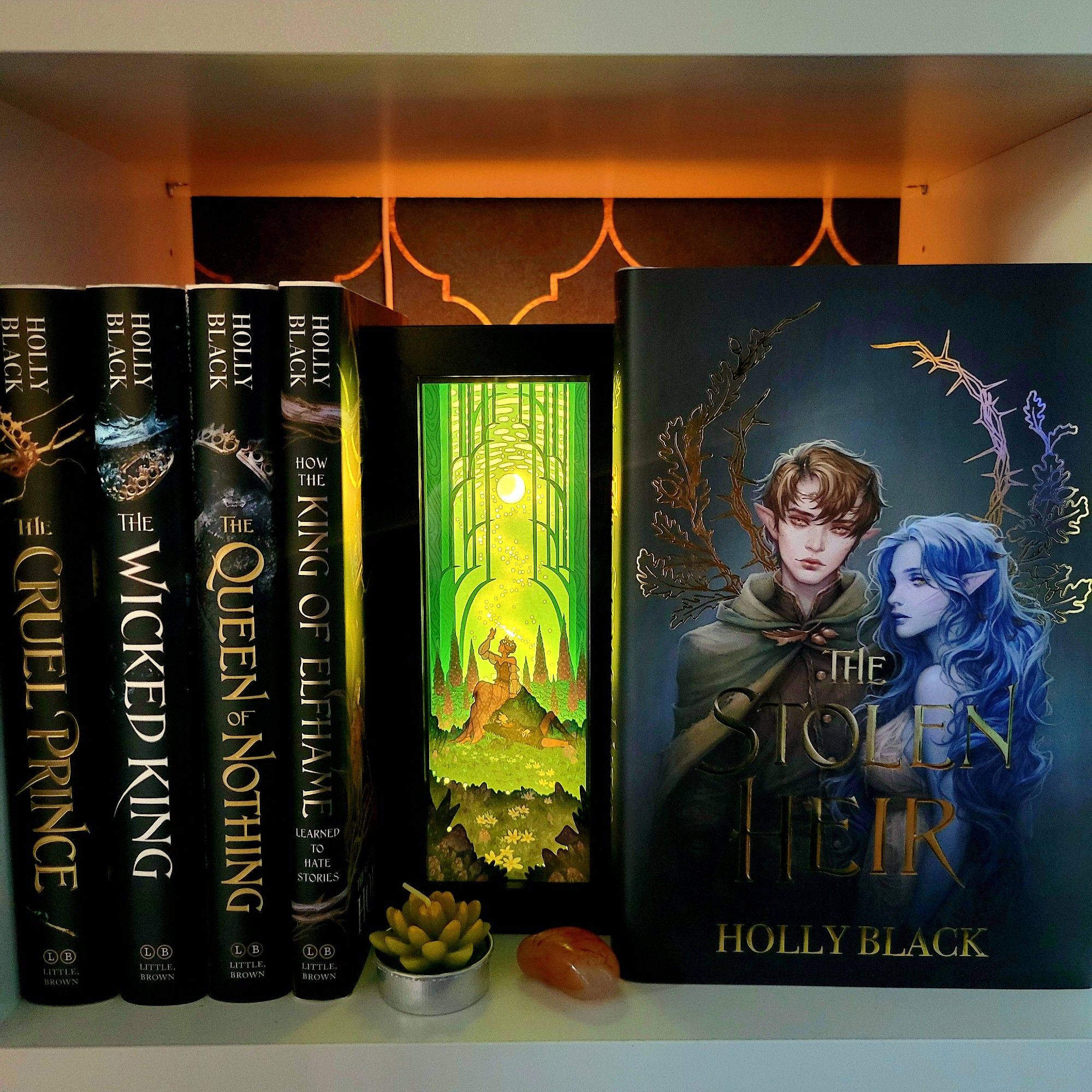 Photo of white bookshelves. Center frame is a lit up box with a scene of Jude and Carden stargazing. Left of the light is a collection of black hardcovers of The Folk of the Air series. To the right is a front-facing hardcover of The Stolen Heir.