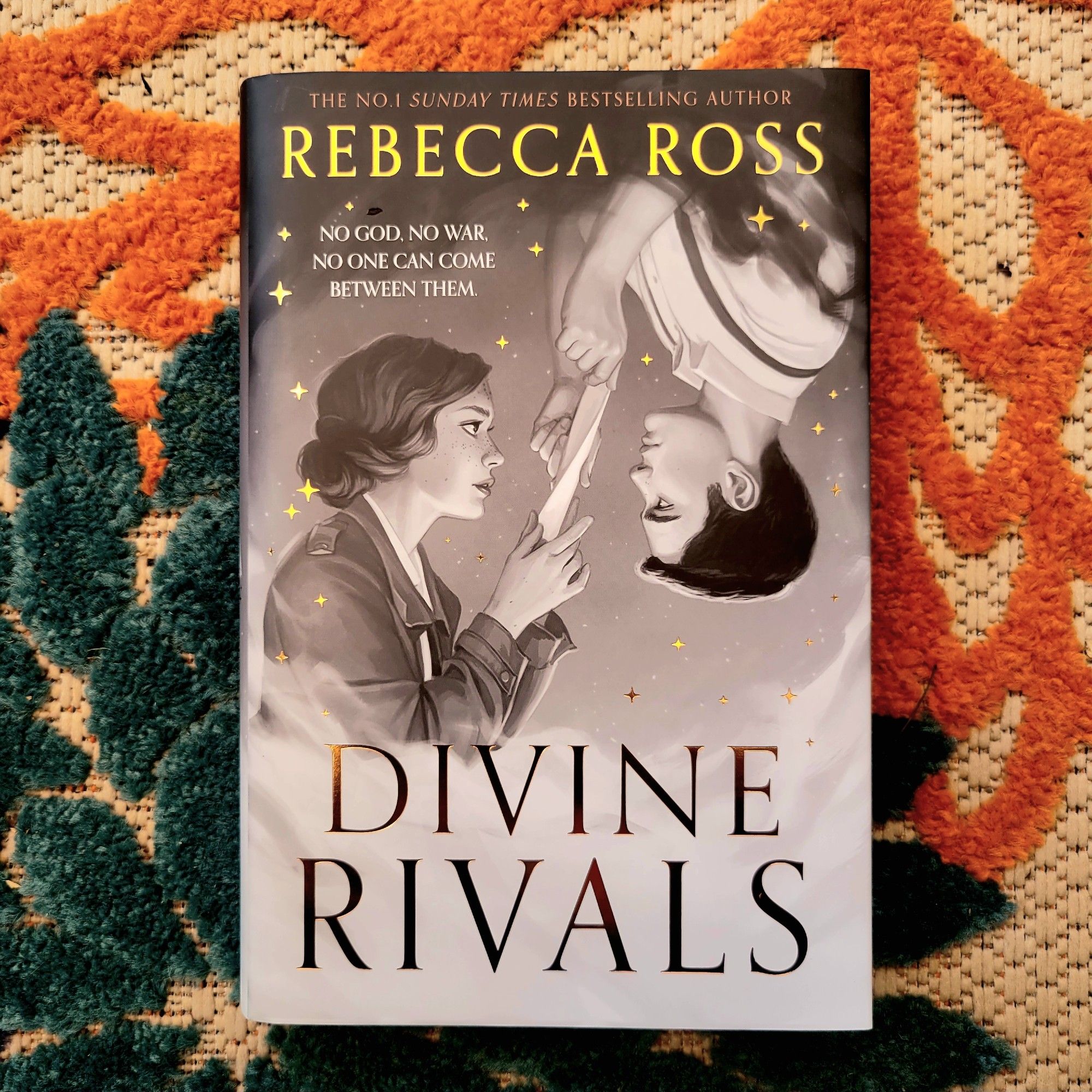 Hardcover book of Divine Rivals by Rebecca Ross, Fairyloot edition, black and white art of a woman and a man in old-fashioned dress, facing each other, holding the same letter between them. Book is set on a background floral pattern of beige, orange, and teal.