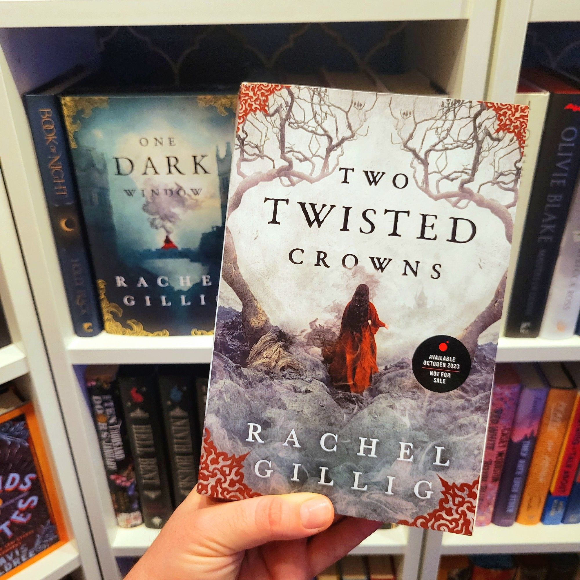 Hand holding an ARC of Two Twisted Crowns by Rachel Gillig. Cover shows a woman with dark hair and a red dress facing away, with two twisted trees on either side. In the background are bookshelves, including One Dark Window by Rachel Gillig.