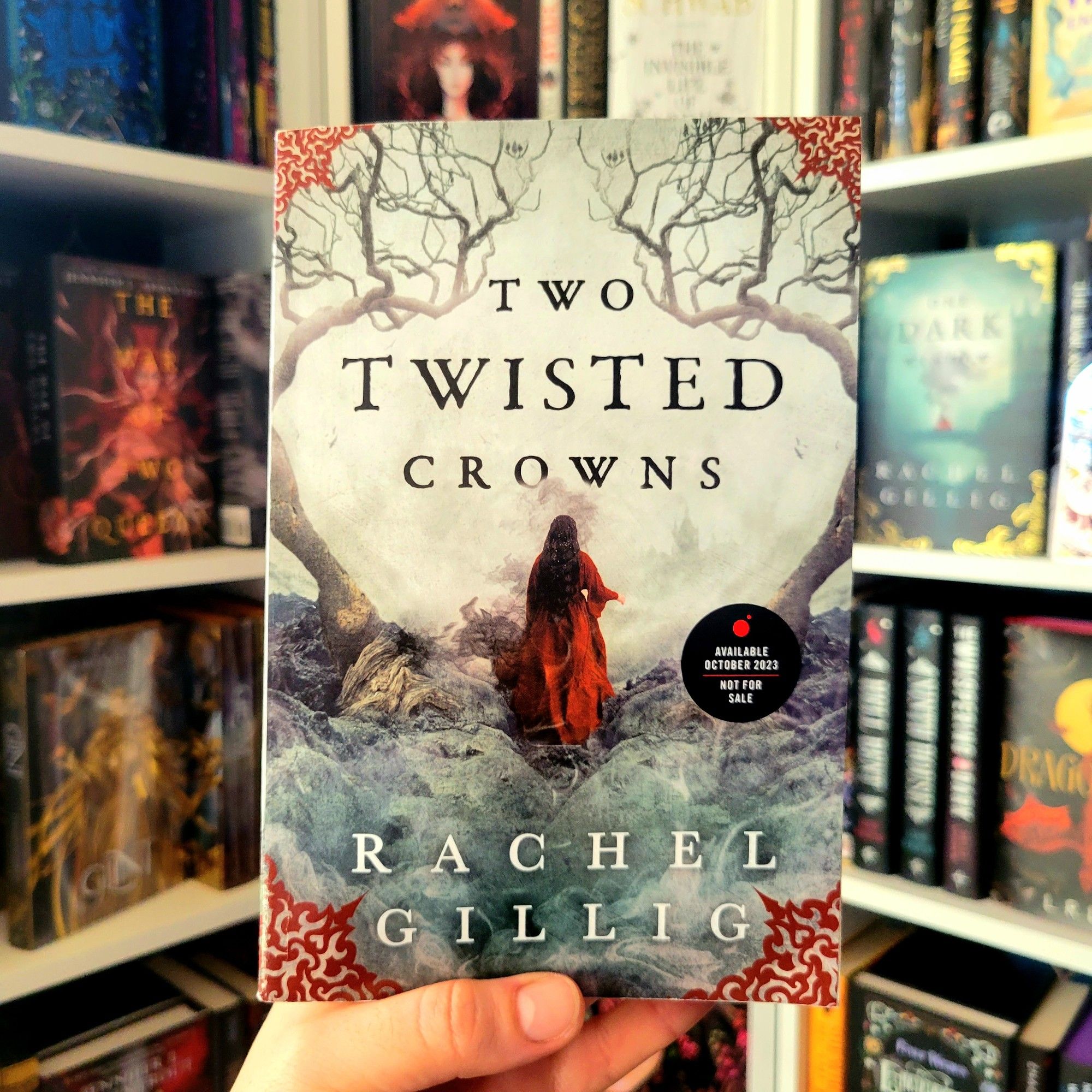 Hand holding an ARC of Two Twisted Crowns by Rachel Gillig. Cover shows a woman with dark hair and a red dress facing away, with two twisted trees on either side. In the background are bookshelves, including One Dark Window by Rachel Gillig.