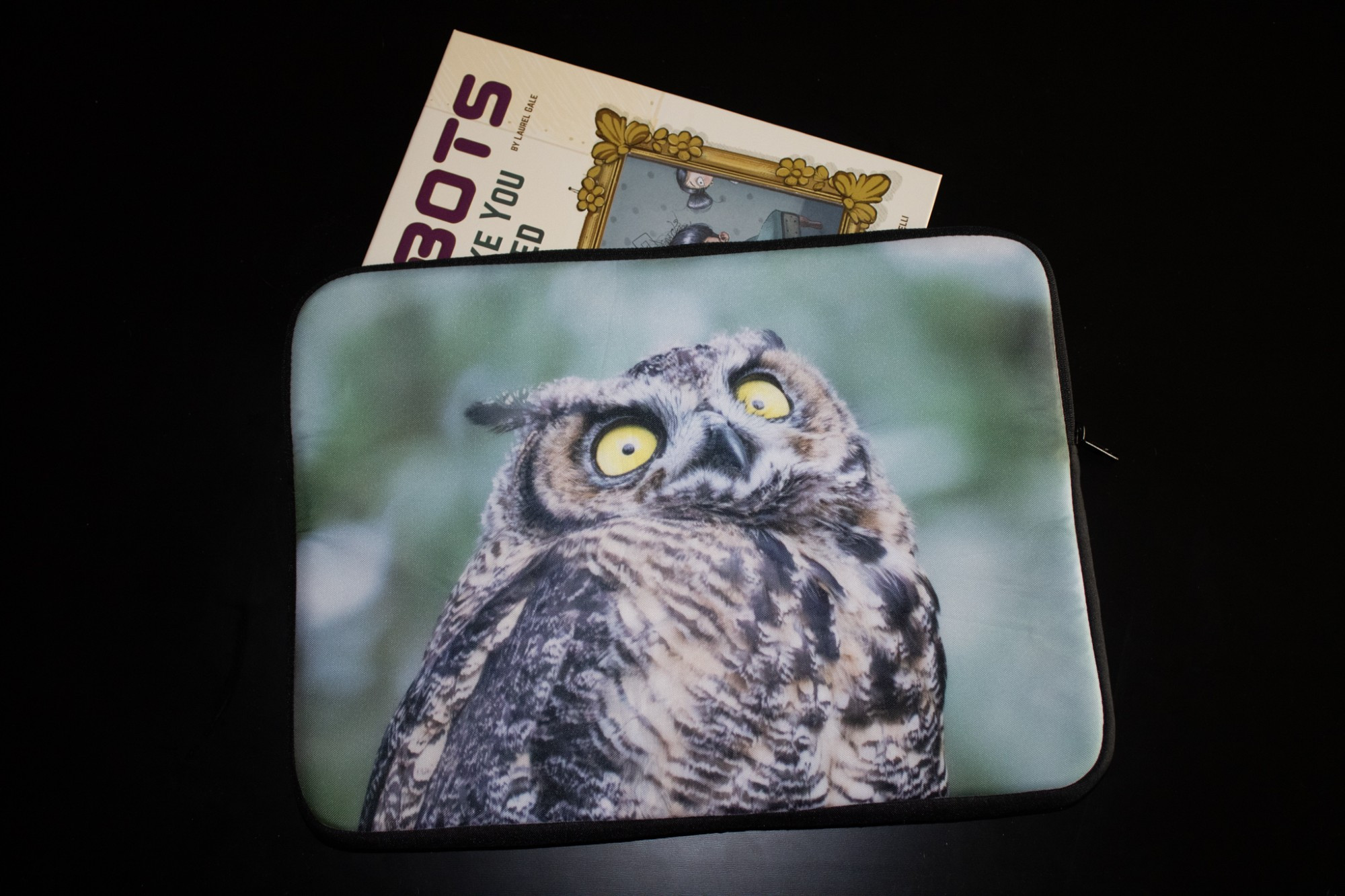 A laptop sleeve featuring a young great horned owl with a wide-eyed expression, holding a copy of the picture book ROBOTS DON'T MAKE YOU GO TO BED.