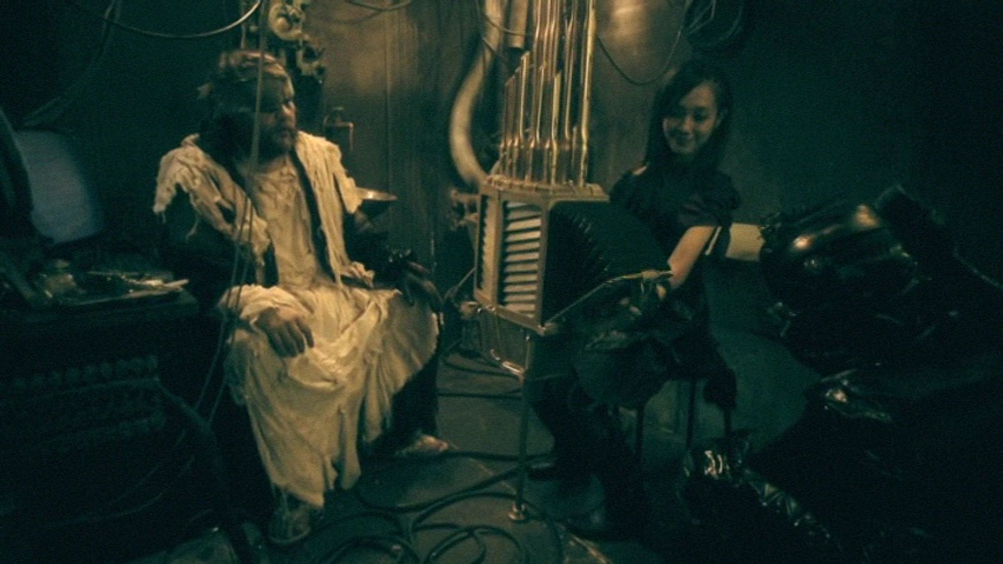 Two ragged figures, a man in white and a black leather clad cyborg, sit in a cyberpunked basement laboratory, watching the black-clad woman play a large musical instrument that looks like a cross between an accordion and a pipe organ