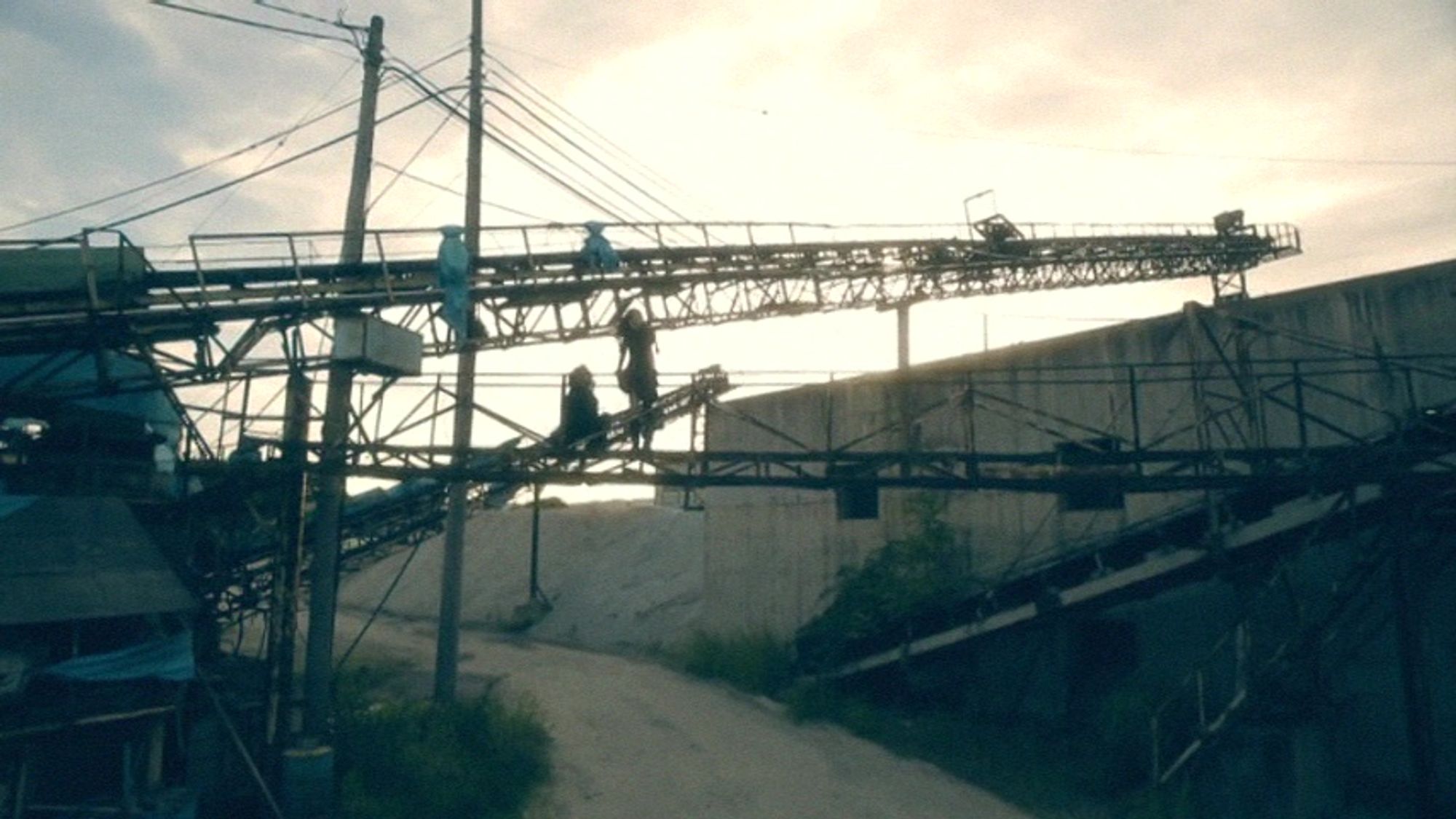 Two human figures in sunset silhouette stand on a crane in an abandoned industrial location.