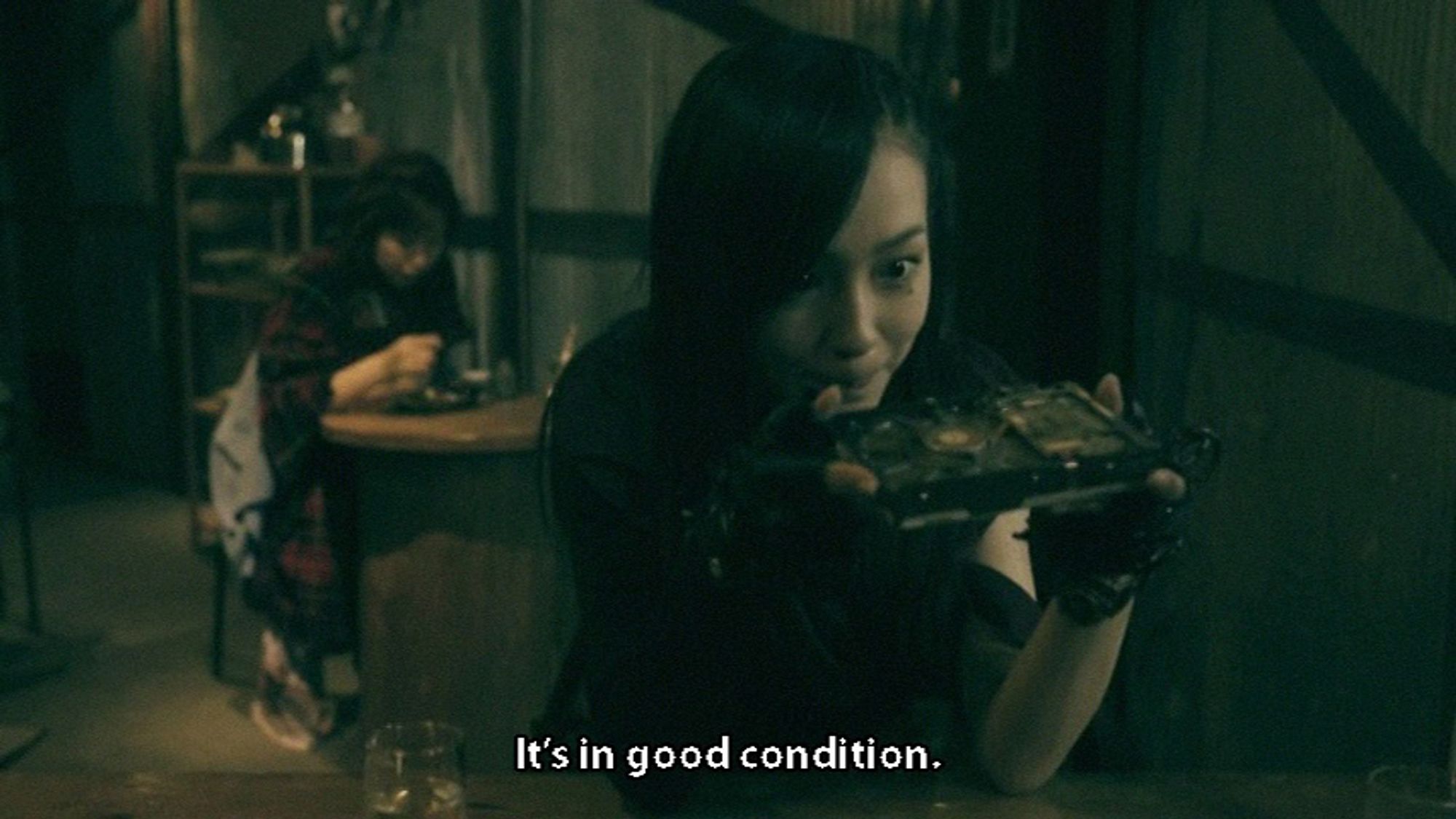 Cyberpunked-out woman in black sits at a table in a decrepit bar-like location, examining a present-day hard drive. Subtitle: It's in good condition.