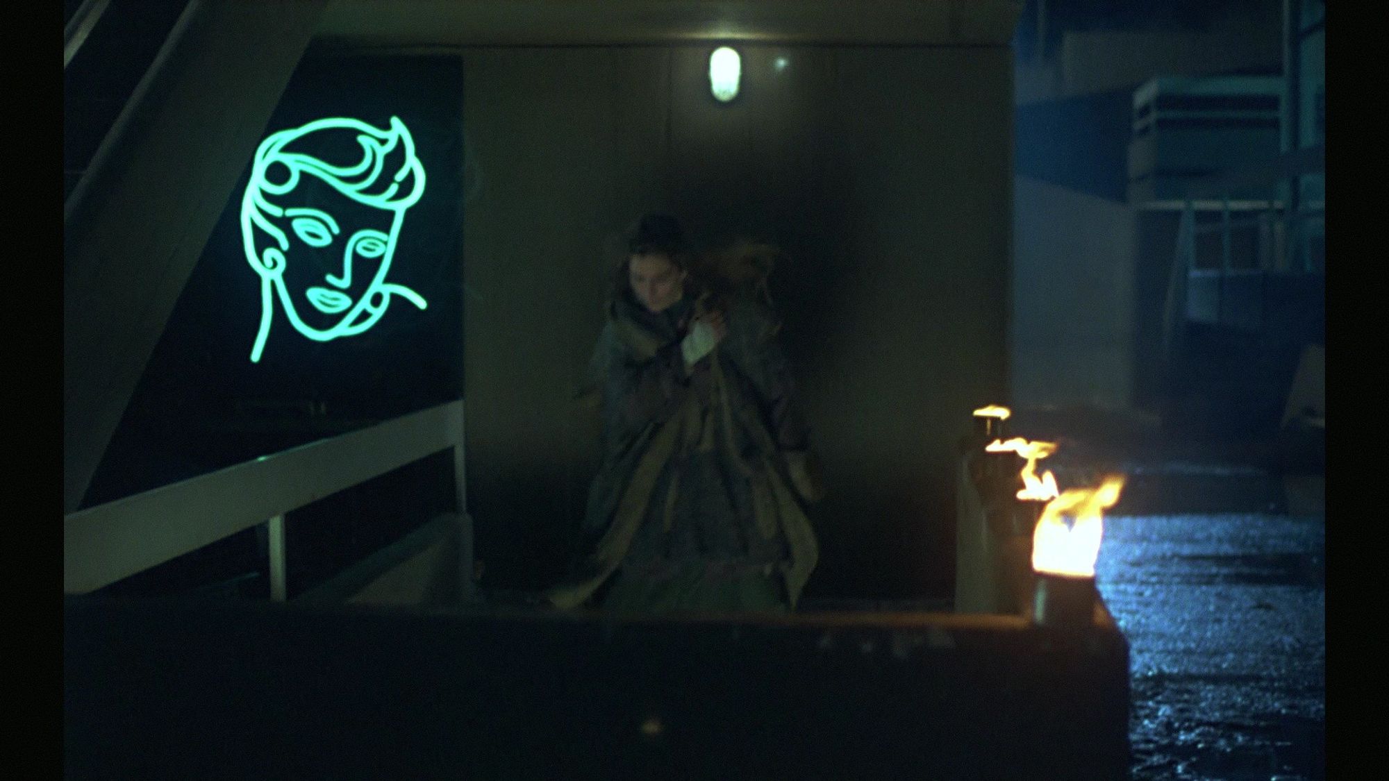 Screenshot from Morning Patrol. A woman descends a staircase in a rainy, grotty concrete environment as trash burns to light the way. A big neon sign (replica of a famous one in LA IIRC) hangs nearby.