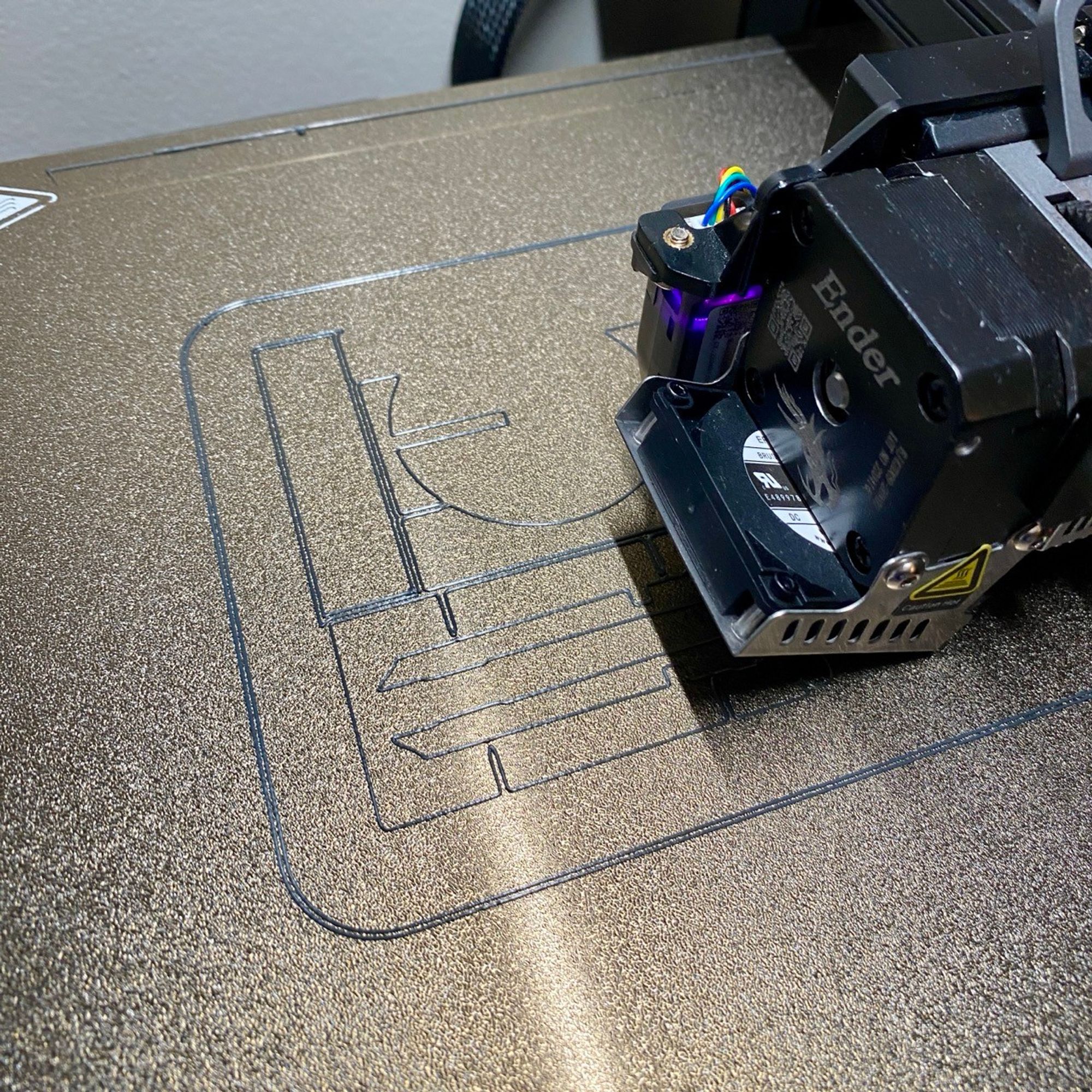 Gold 3D print bed and black Ender sprite hot-end with silver grey filament forming rectangular kit card. Only a few lines if filament are visible but you can see 2 rectangular sections and one large round saucer shape.  